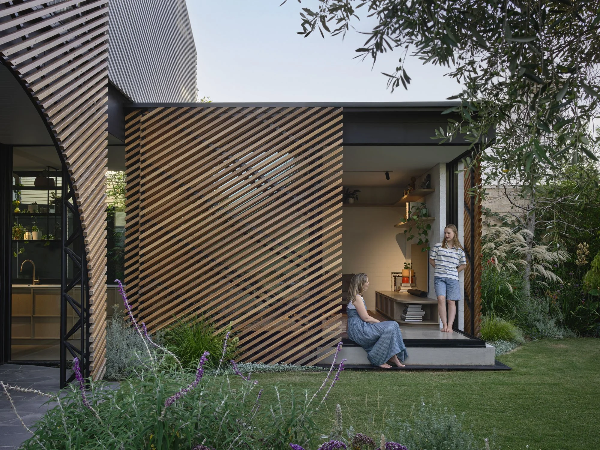Modern house with wooden slat exterior and large glass windows, surrounded by garden greenery and purple flowers, with two women inside, one sitting and one standing.