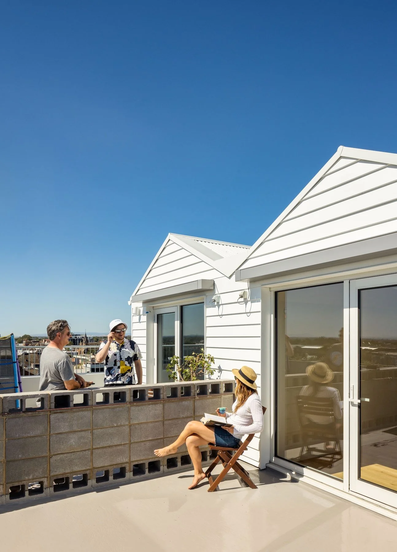 Group of four people on a rooftop balcony engaging in conversation, with one woman sitting on a chair holding a cup and a notebook, and the others standing near a railing, with buildings and a blue sky in the background.