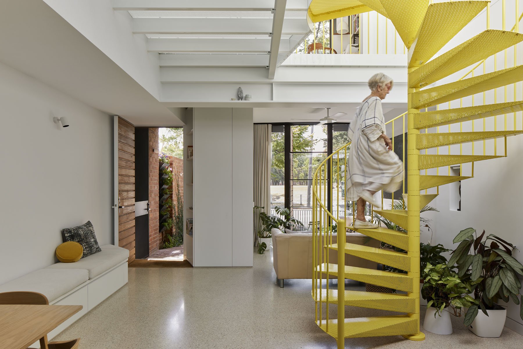 An elderly woman with gray hair, wearing a loose white dress, is ascending a vibrant yellow spiral staircase inside a modern, well-lit home. The interior features white walls, large windows with curtains, and various plants. The space includes a sitting area with a white bench, a small wooden table, and decorative pillows.