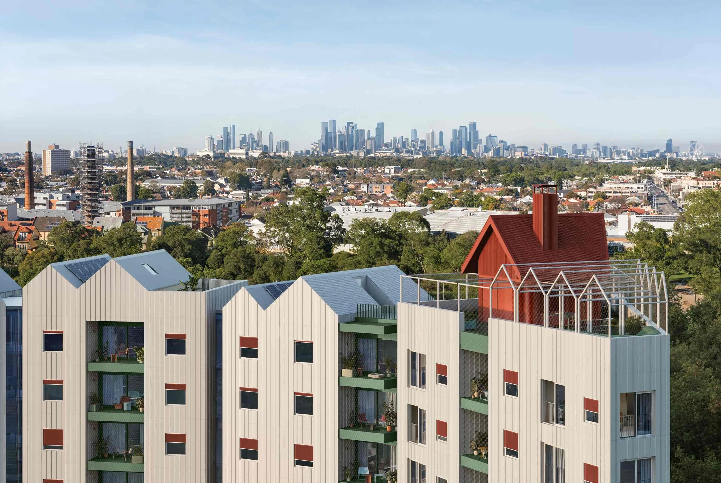 A view of an urban neighborhood with modern apartments and lush green trees, with a city skyline of tall skyscrapers in the background against a clear blue sky.