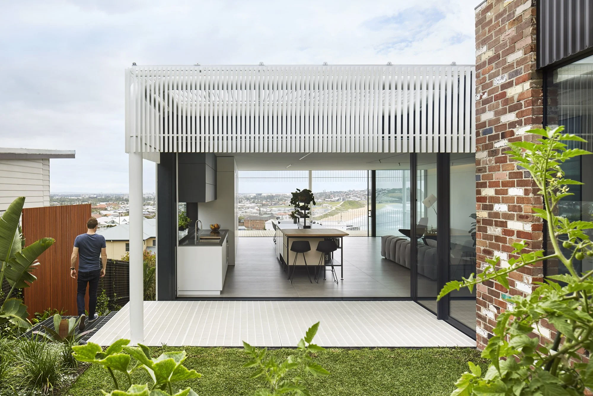 A modern rooftop terrace featuring a covered outdoor kitchen and dining area with a view of a cityscape in the background. The space is decorated with plants and has a sliding glass door leading inside, with a brick wall on the right.