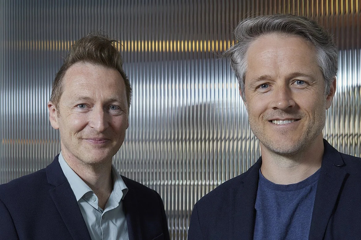 Two men in business attire standing in front of a modern, metallic, ribbed wall, smiling at the camera.