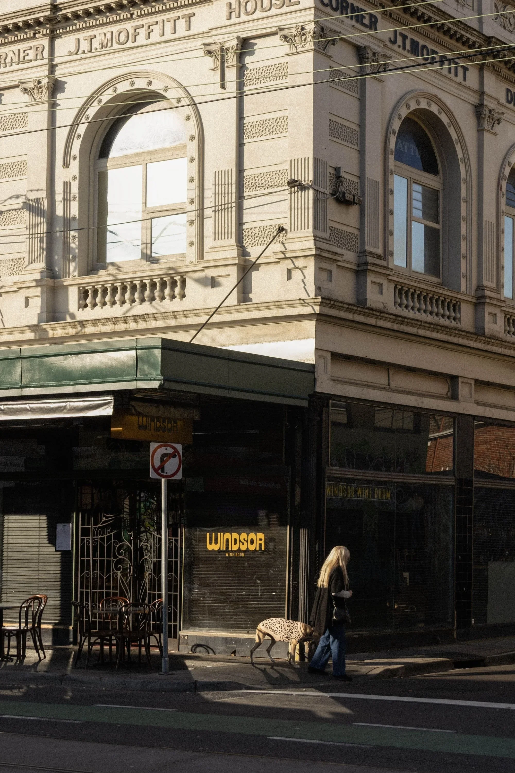 The corner of a historic building with ornate architecture and arched windows, featuring signage for J.T. Moffitt. On the street, there are outdoor tables and chairs, a person walking with a pet dog, and a sign indicating no U-turns.