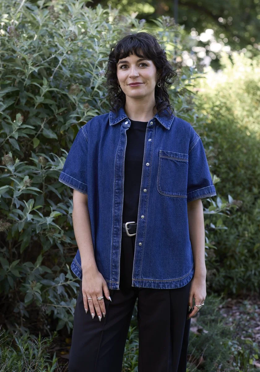 A woman with short curly black hair standing outdoors among green foliage, wearing a dark blue denim shirt over a black top, black pants, silver jewelry, and holding a phone in her right hand.