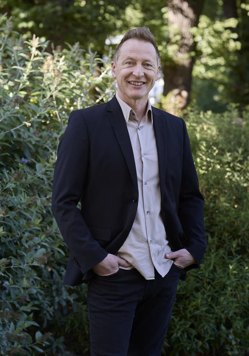 Smiling man in a black jacket and beige shirt standing outdoors among green foliage.