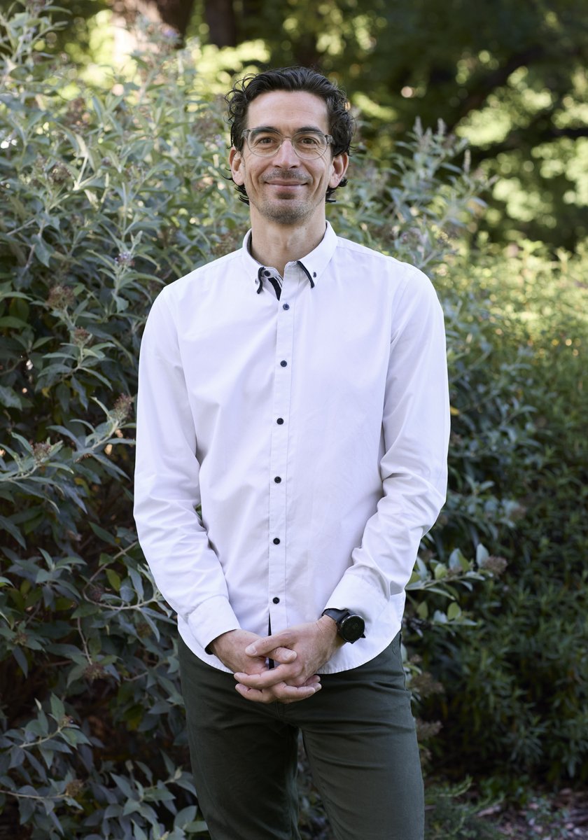 A man wearing glasses and a white button-up shirt stands outdoors in front of green bushes, smiling at the camera.
