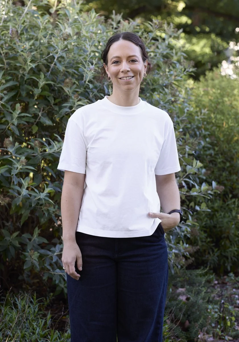 A woman with dark hair, wearing a white t-shirt and dark jeans, standing outdoors in front of green leafy bushes and smiling.