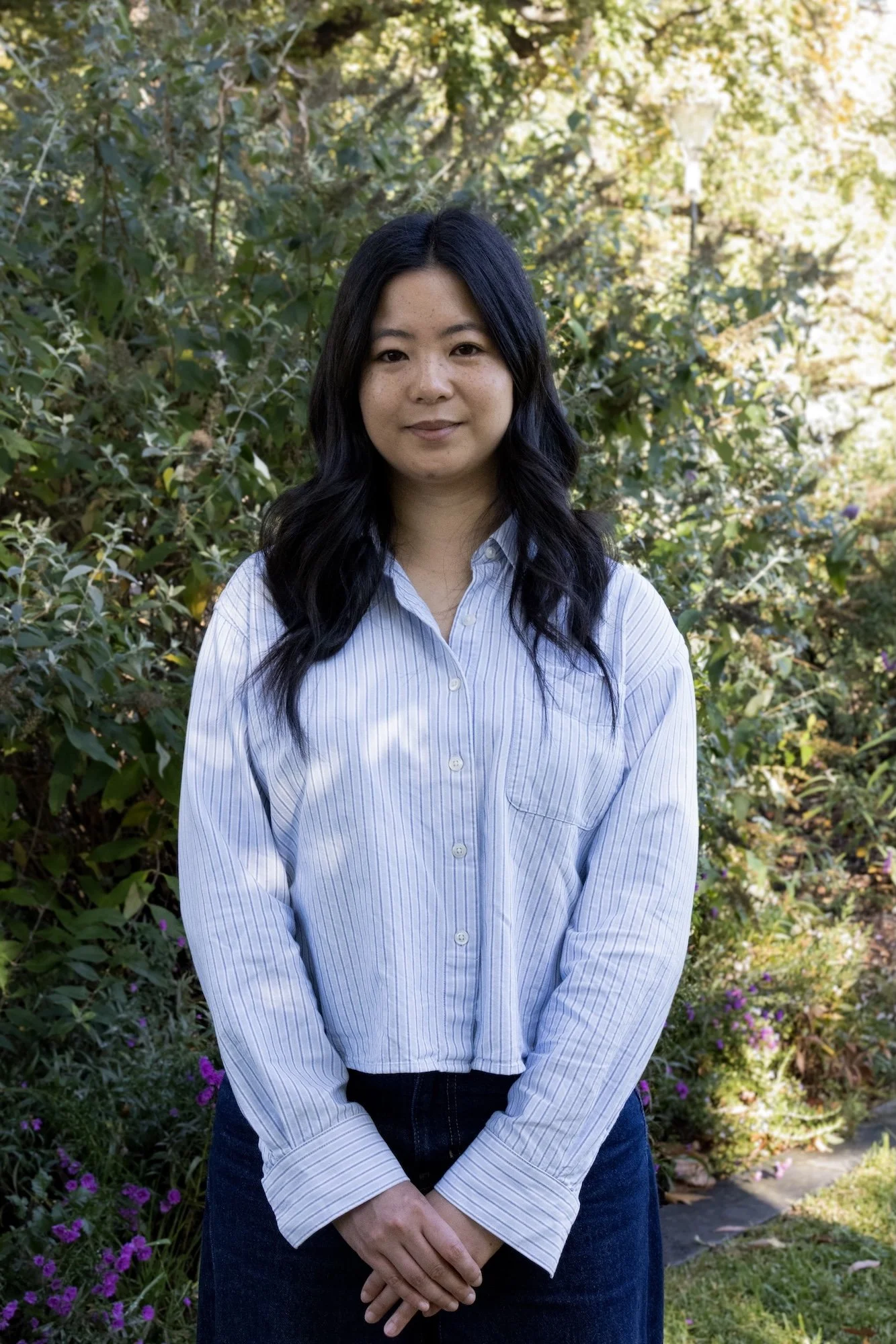 Woman with long dark hair wearing a light blue striped shirt standing outdoors in front of green foliage and purple flowers.
