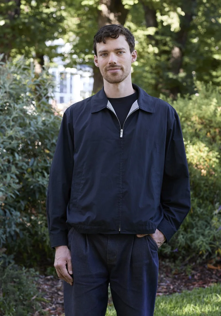 A young man with dark curly hair and a beard standing outdoors in a park with green trees and bushes. He is wearing a black zip-up jacket over a black shirt and has his right hand in his pocket.