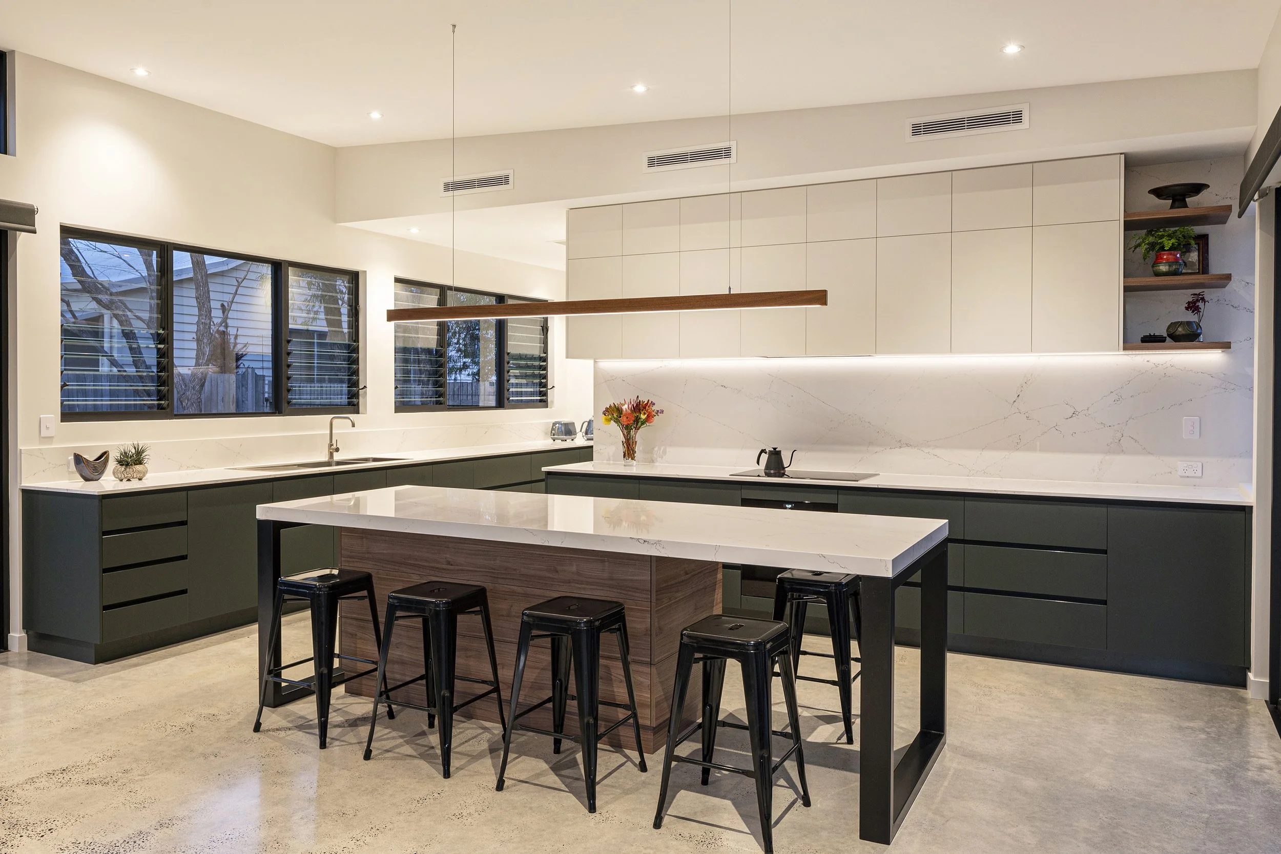 Modern kitchen with white upper cabinets, black lower cabinets, marble backsplash, a large island with a white marble countertop, black bar stools, and a row of windows with venetian blinds.