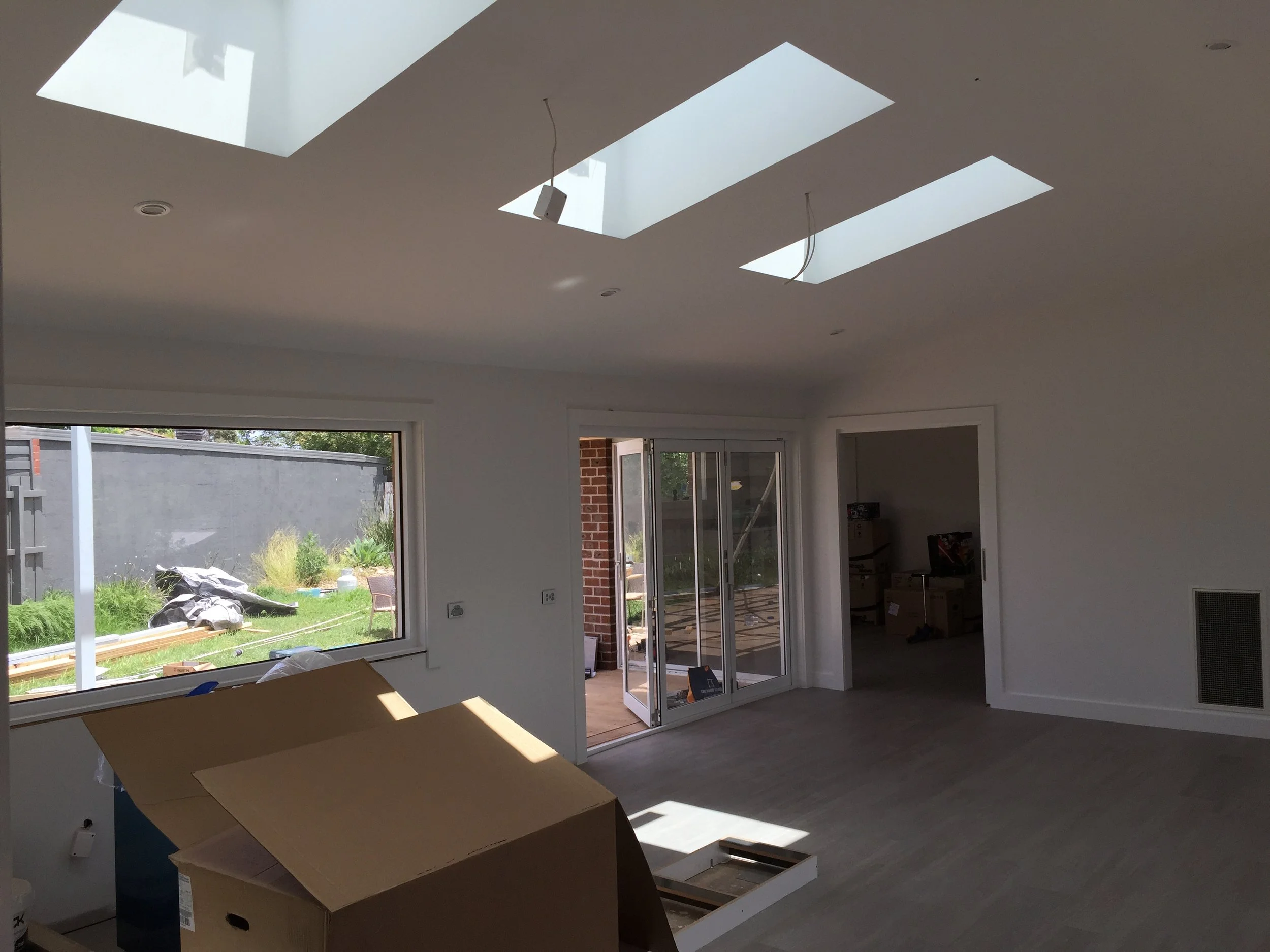 Interior of a house under construction with large skylight windows in the ceiling, sliding glass door leading to an outdoor yard, and construction materials and tools scattered inside.