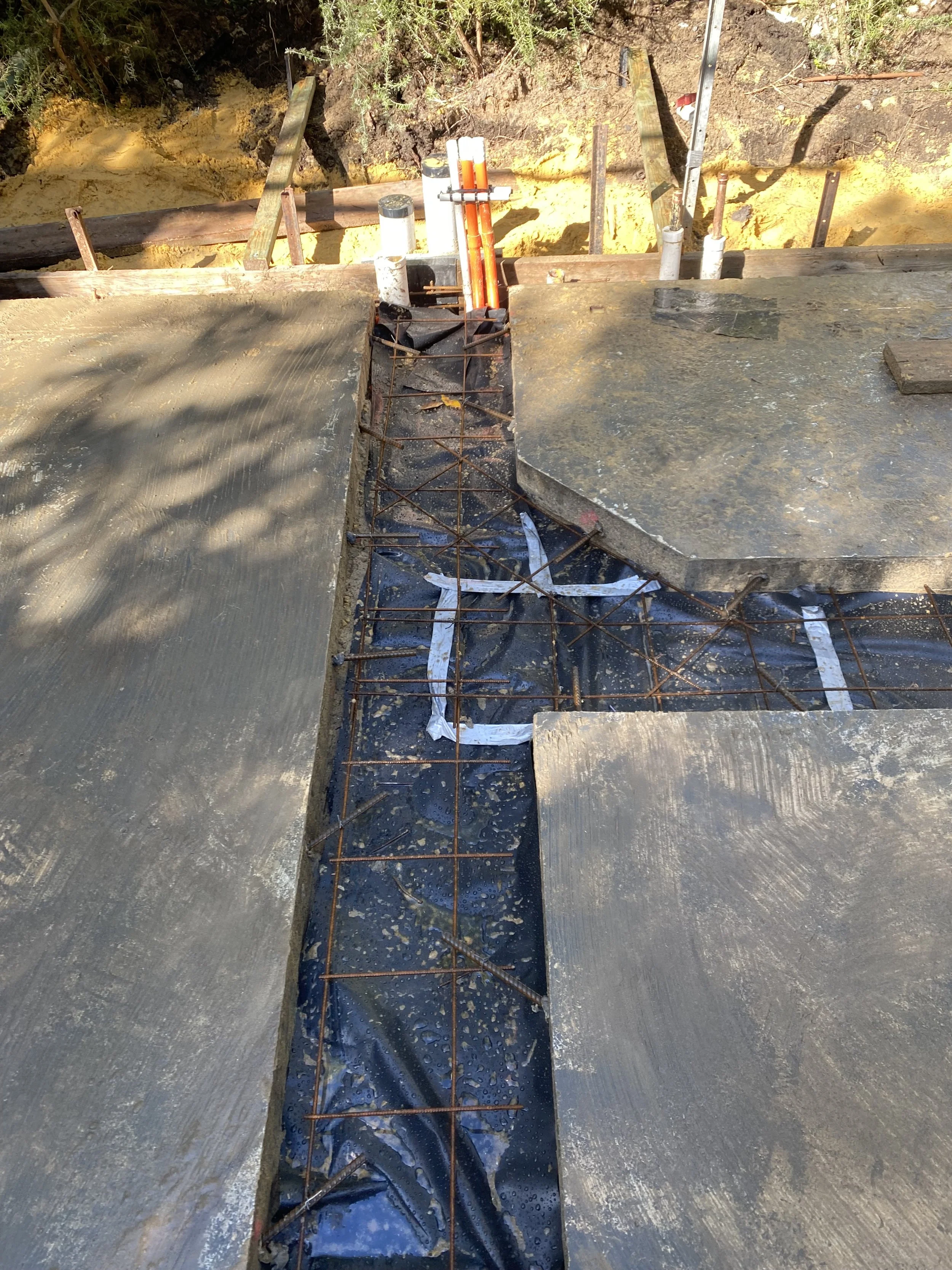 Construction site showing a partially built concrete driveway or pathway with rebar framework, some pipes, and wooden formwork, surrounded by dirt and soil.