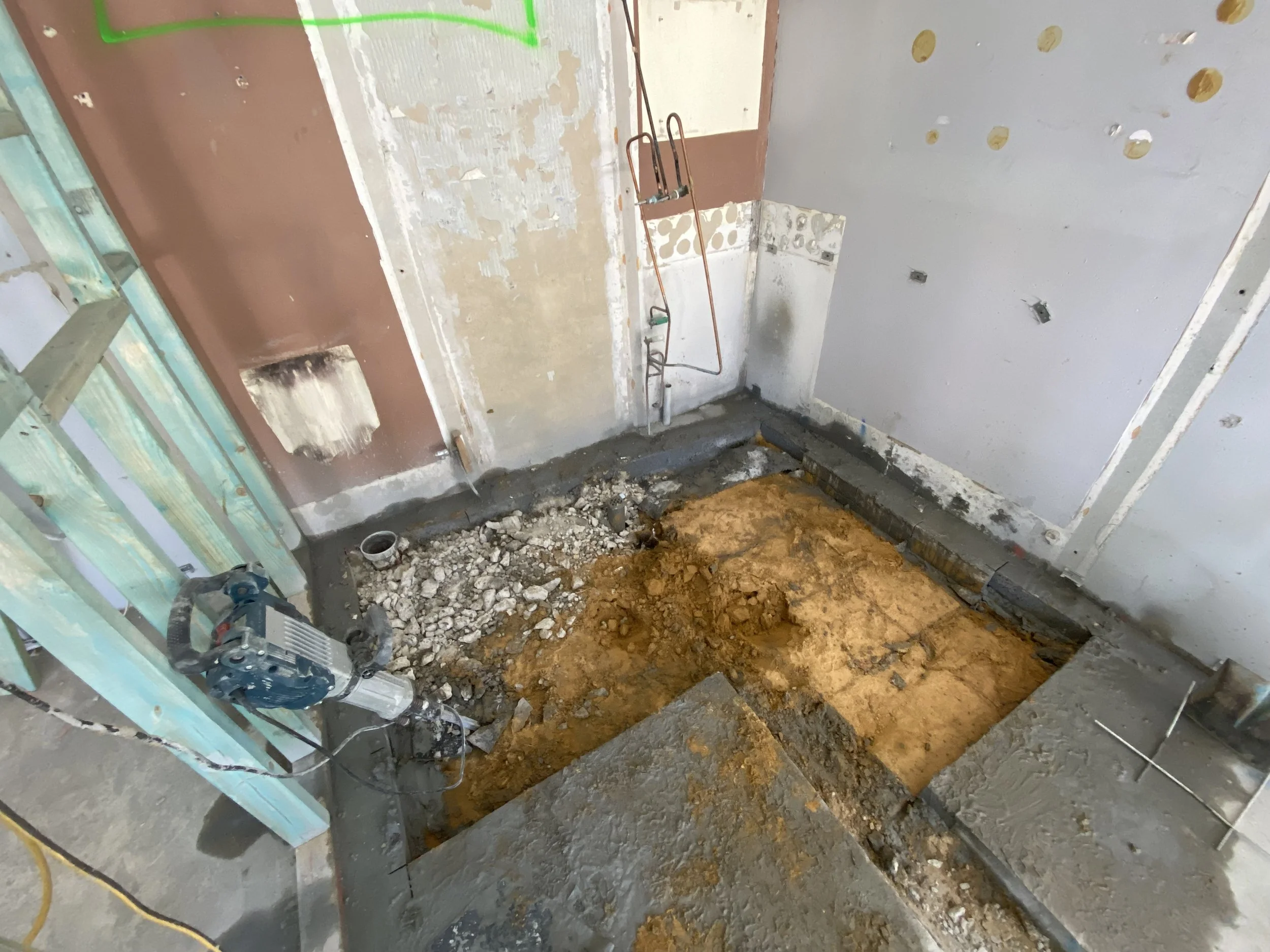 A room under construction with an exposed floor filled with dirt and debris. The walls are partially finished with drywall in some areas and there are pipes and electrical wiring visible. Construction tools and materials are scattered around.