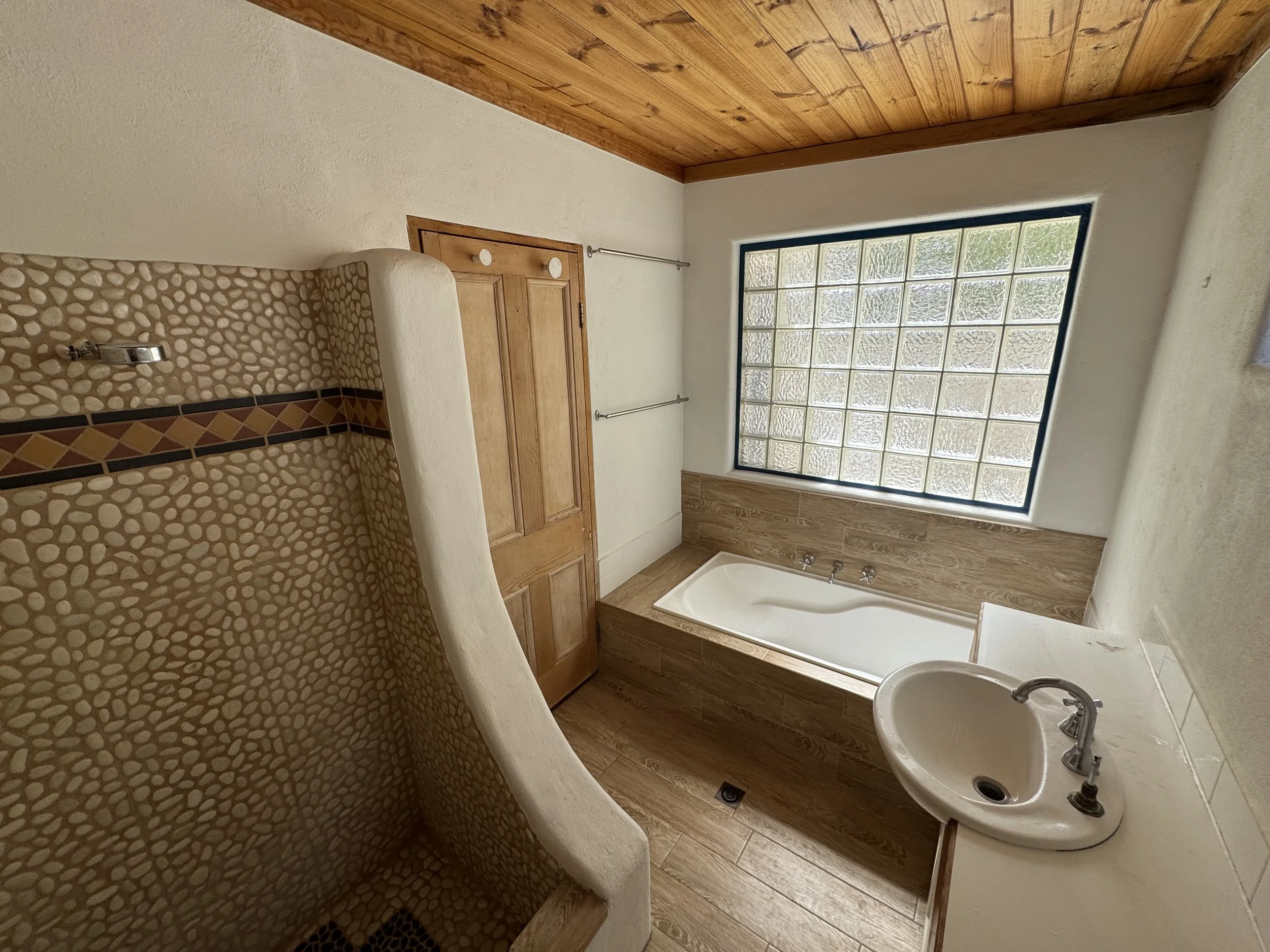 A bathroom with a corner shower with pebble tile walls, a wooden door, a large window made of glass blocks, a bathtub, a sink with faucets, and wood accents on the ceiling and floor.