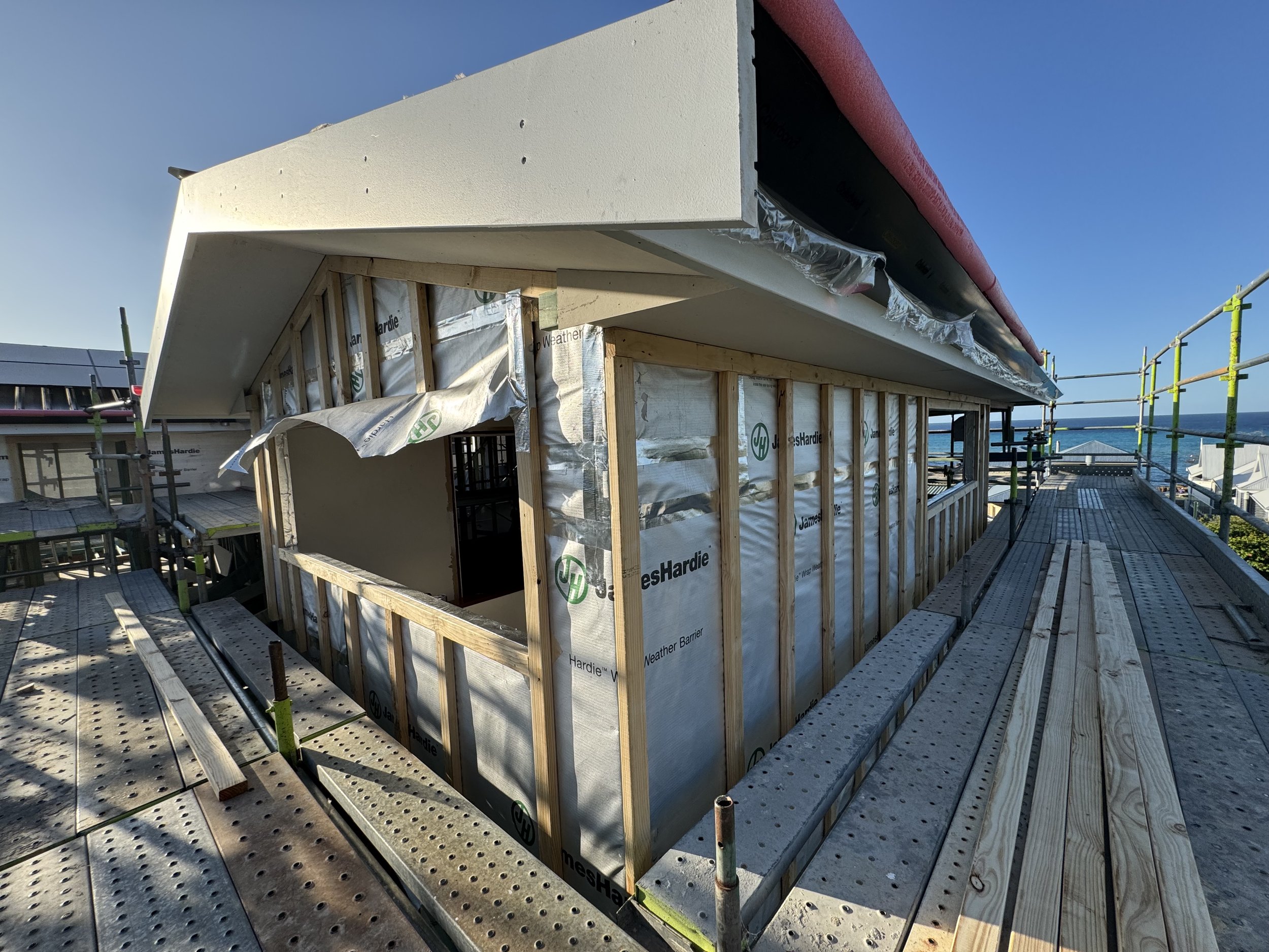 Construction site on a rooftop with scaffolding and the framing of a building under construction, with the ocean and a clear blue sky in the background.