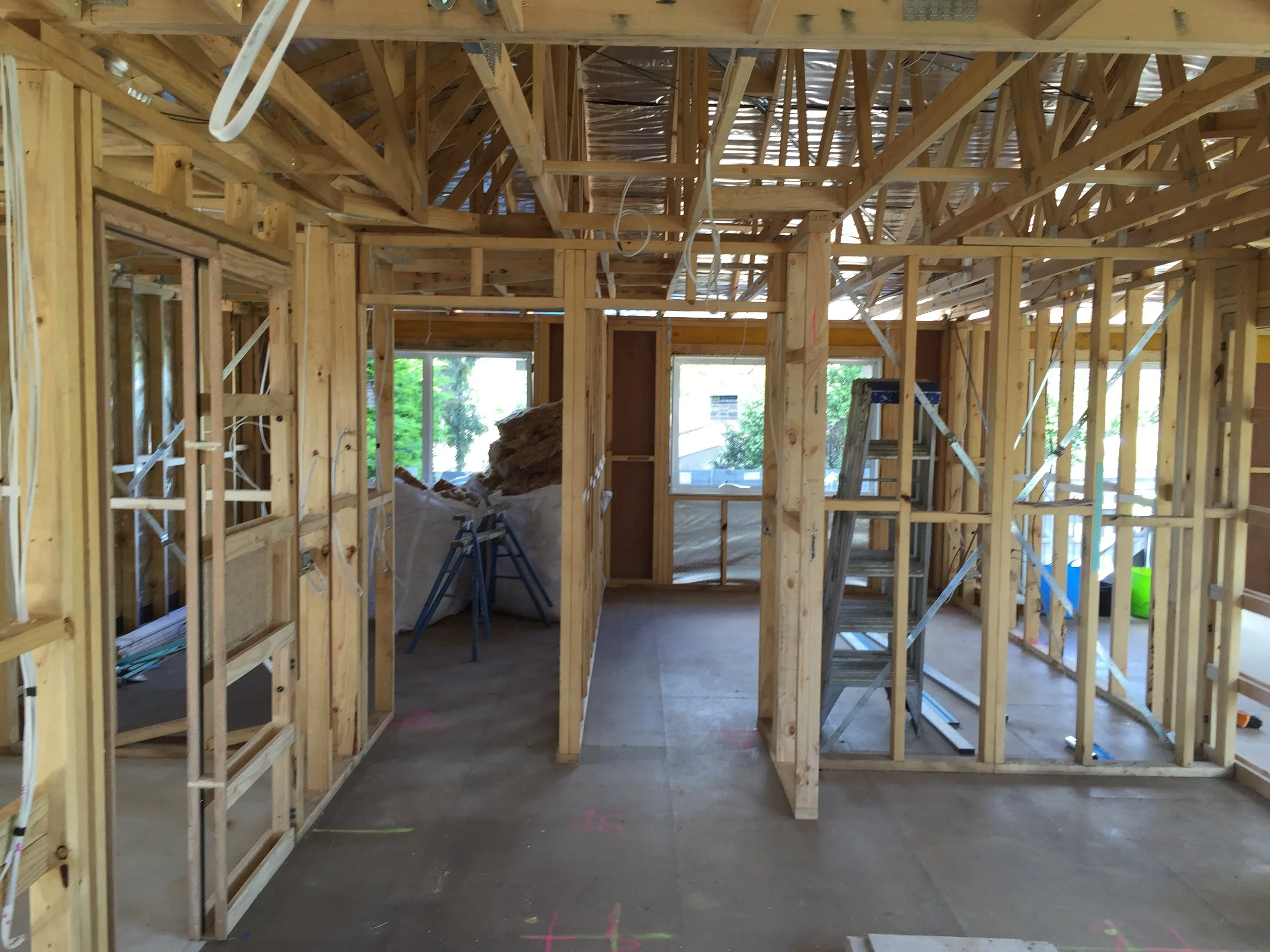 Interior view of a house under construction showing wooden framing for walls and ceiling, with some windows and construction tools present.