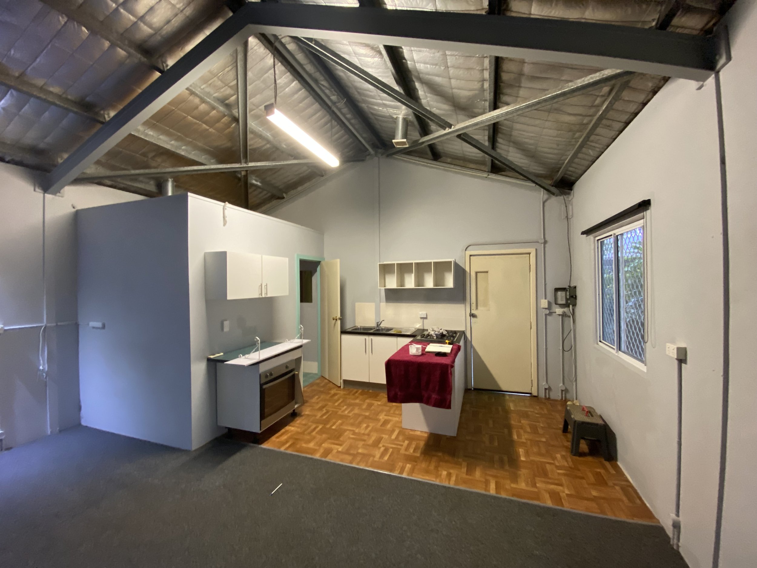 A room with partially finished interior construction, showing exposed metal roofing and insulation on the ceiling, a window with a security screen, cabinets, and a table with a red cloth.