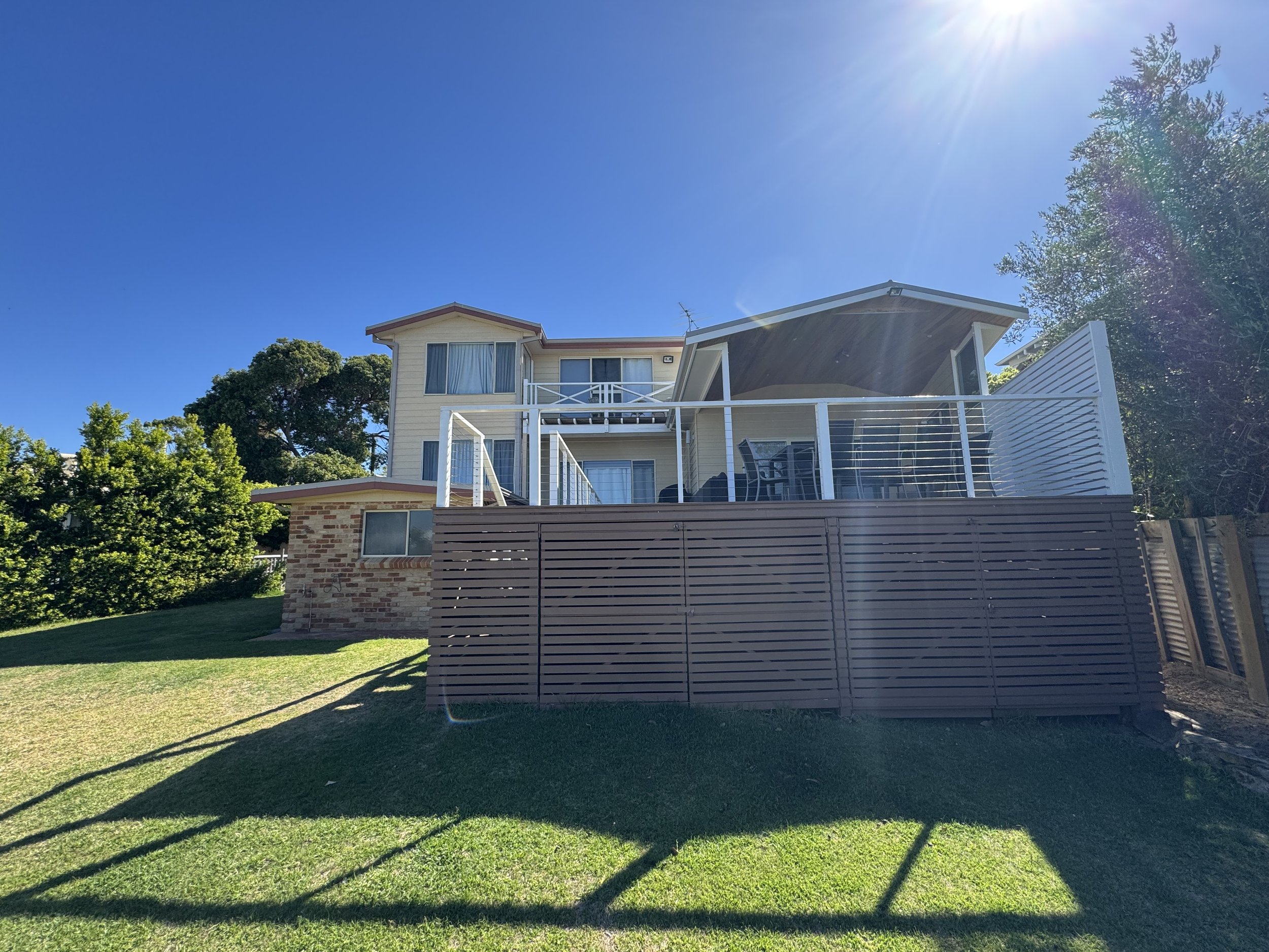 Two-story house with a balcony and large windows, surrounded by green trees and a well-maintained lawn, under a clear blue sky with the sun shining bright.