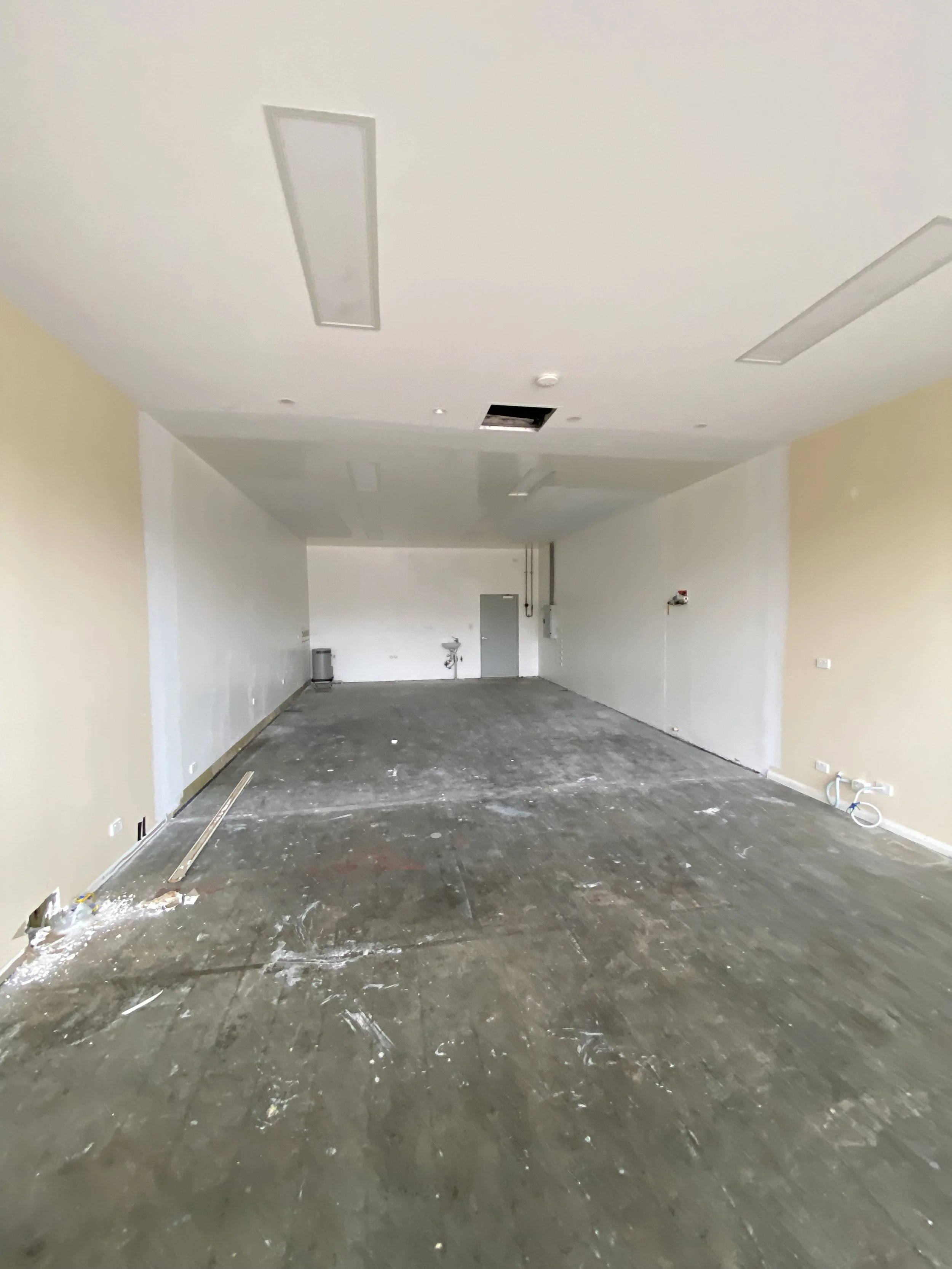 Empty room under renovation with a bare concrete floor, electrical wiring, wall outlets, and ceiling lights, some panels missing from the ceiling.