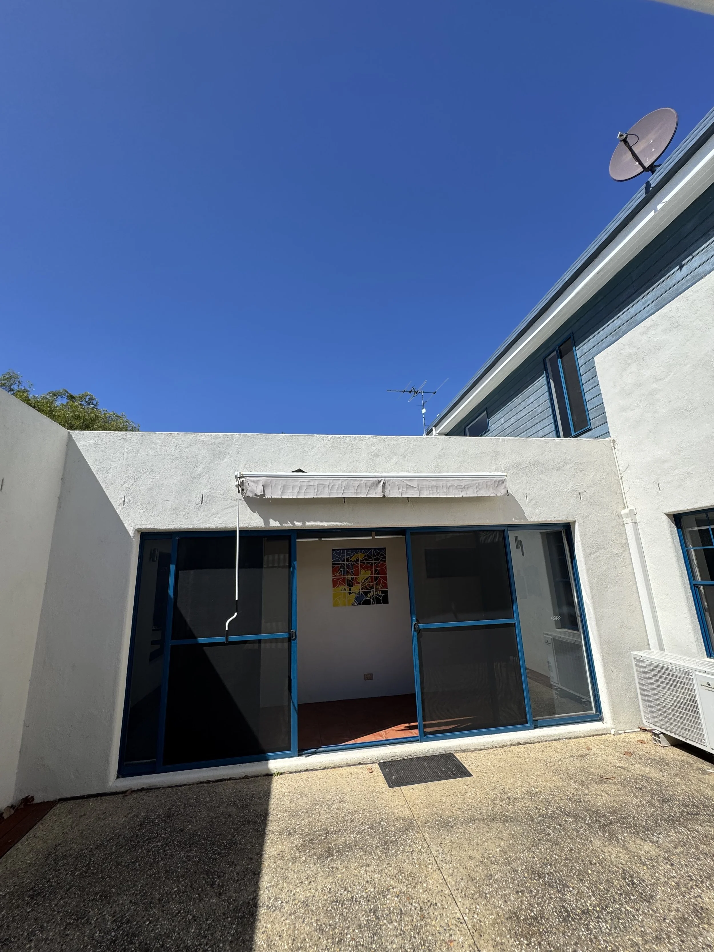 Outside view of a house with a sliding door and an awning, with a border wall and an open space in the foreground, blue sky in the background.