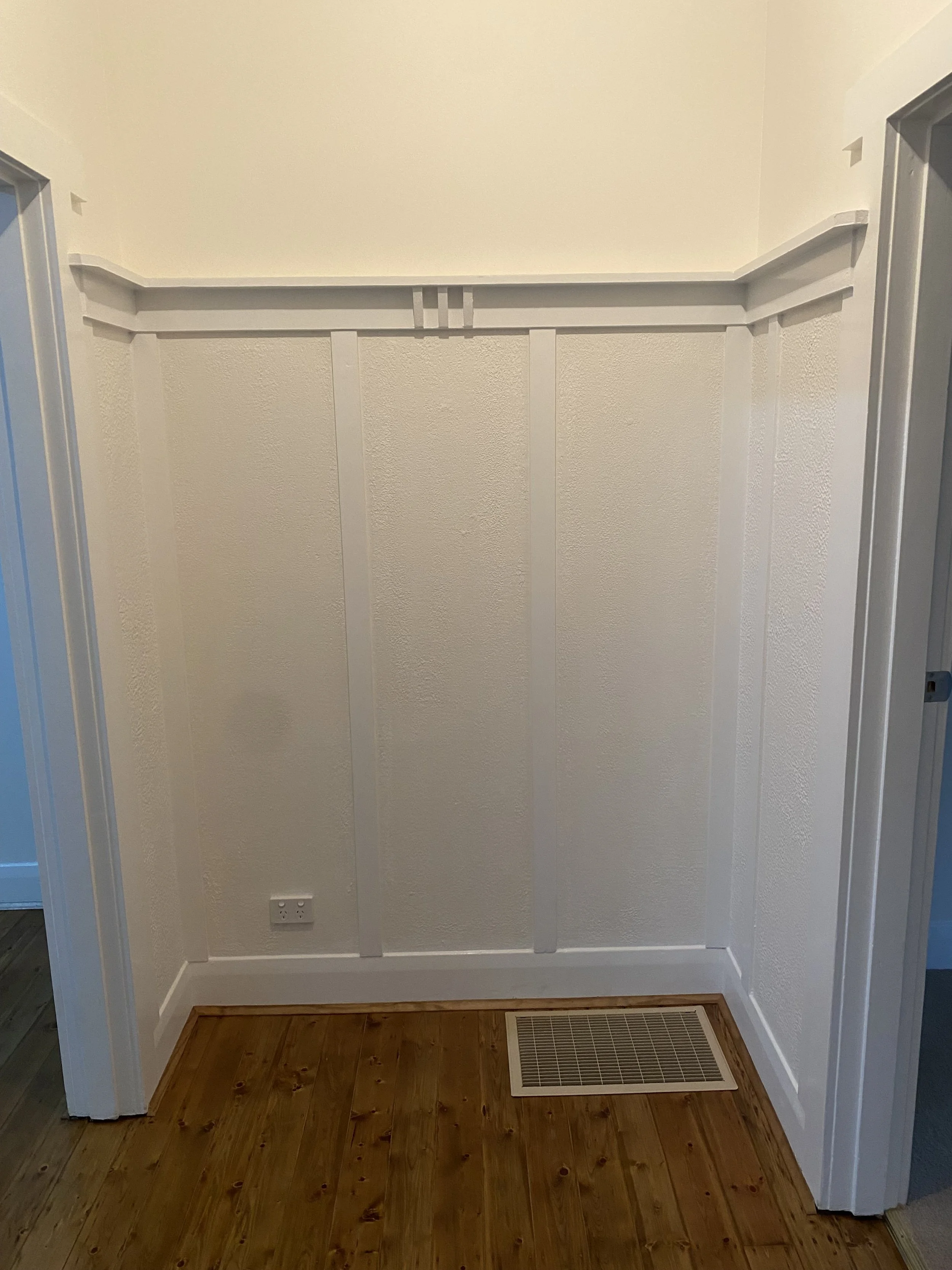 Empty corner of a room with white walls, wooden floor, an electrical outlet on the wall, and a vent on the floor.
