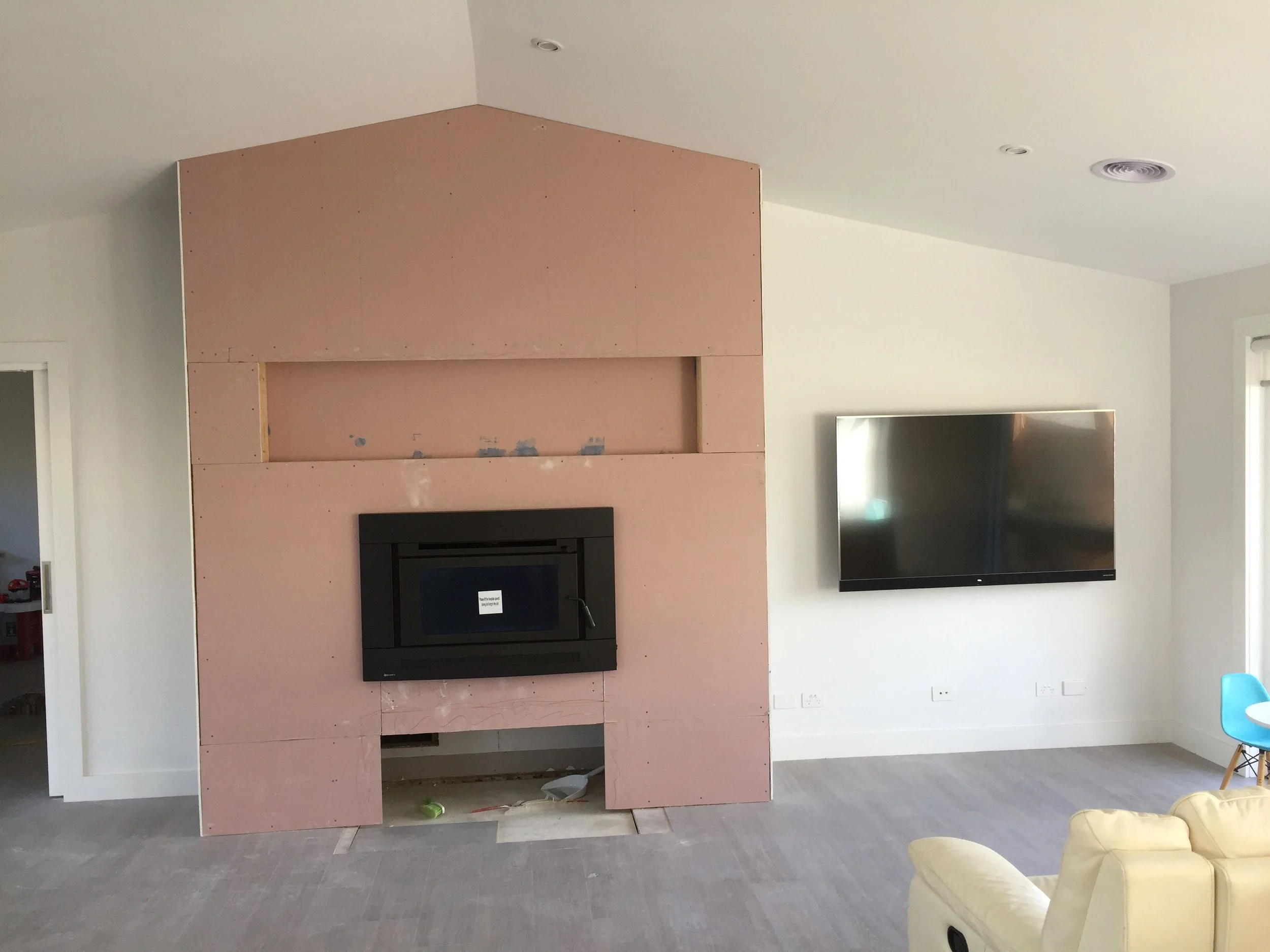 Living room with an unfinished pink drywall wall, a black fireplace, a flat-screen TV, a gray tile floor, a partially visible yellow sofa, and a blue chair.