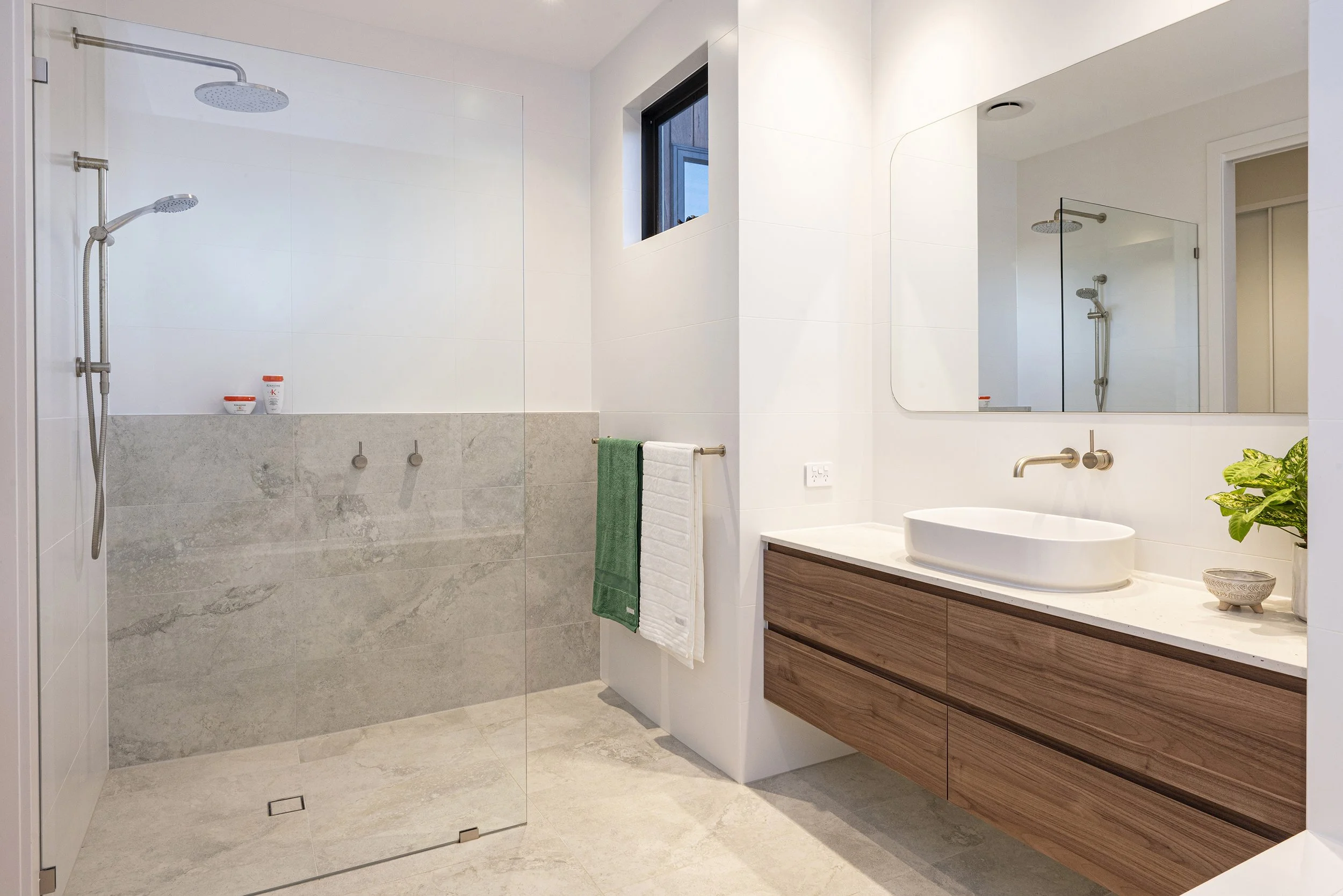 Modern bathroom with a walk-in shower featuring two showerheads, a large mirror, a vessel sink on a wooden vanity, a small plant, and a towel hanging on a rail. The bathroom has neutral-colored tiles and a high window bringing natural light.