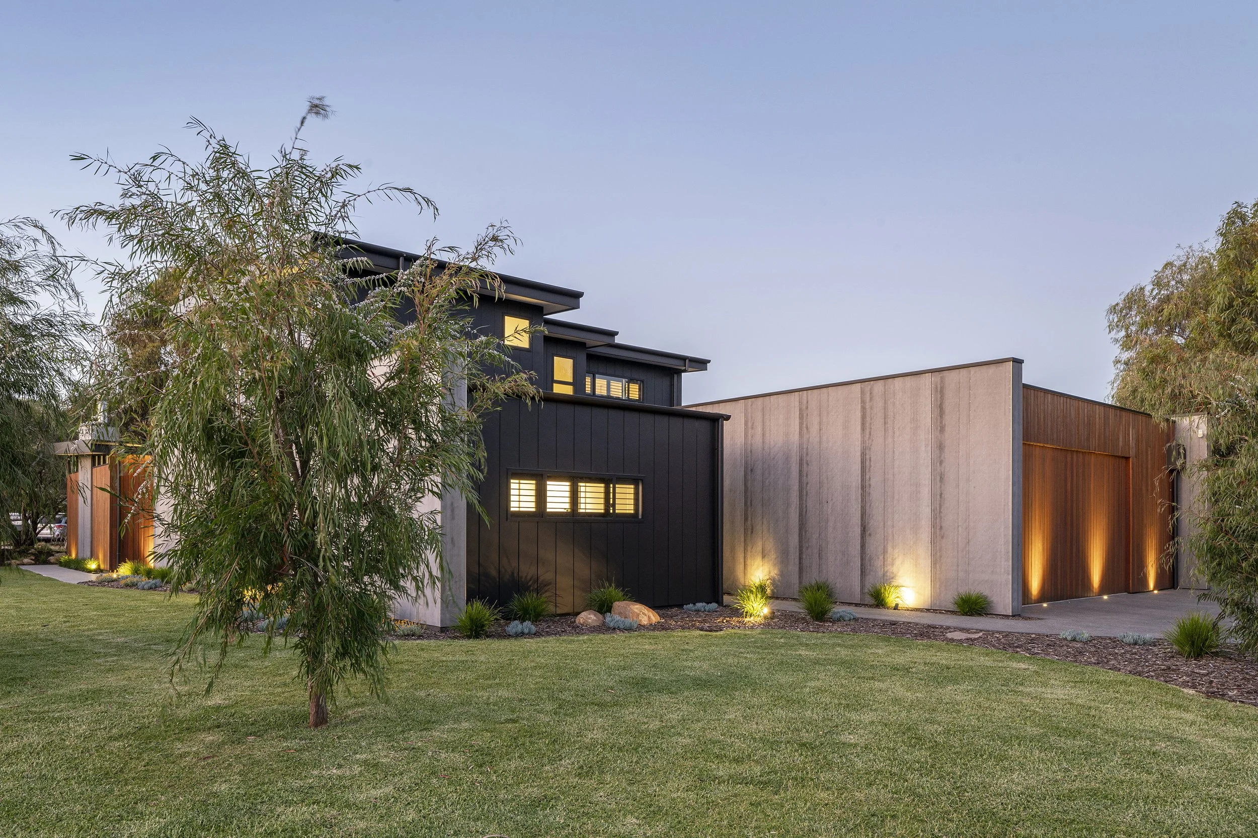 Modern residential house exterior at dusk with illuminated windows, landscaped yard, trees, and external lighting.