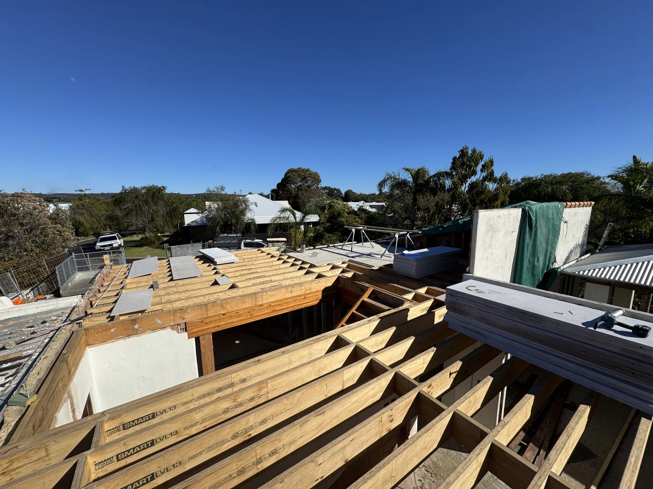 Building under construction with exposed wooden framing and roofing materials, surrounded by trees and residential area.