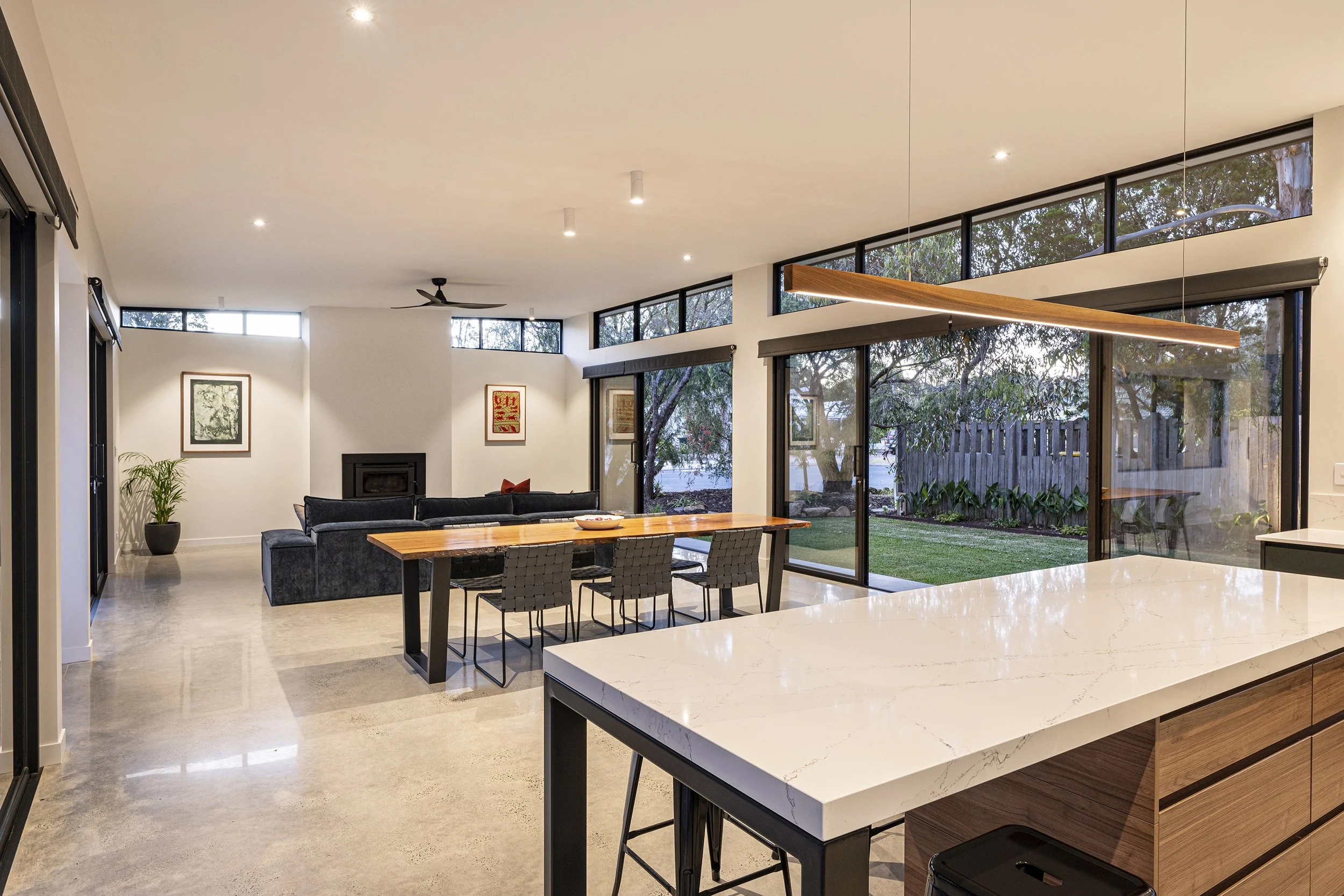 Modern open-concept living room and kitchen with large windows, a black fireplace, gray sofa, wooden dining table, and granite countertops.
