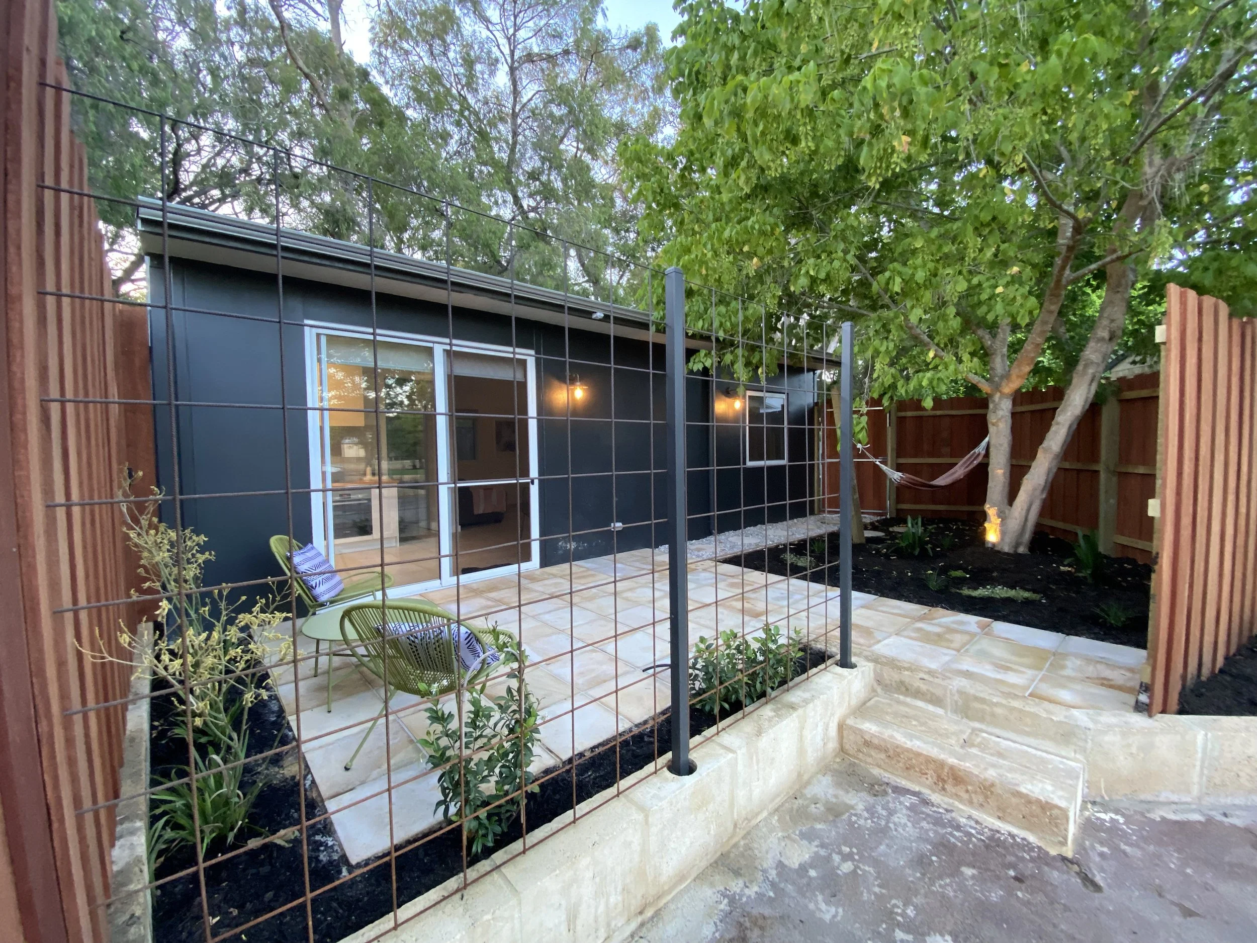 A backyard with a black building with large windows, a small patio with two green outdoor chairs, a garden bed with plants, a tree, a wooden fence, and a wire trellis.