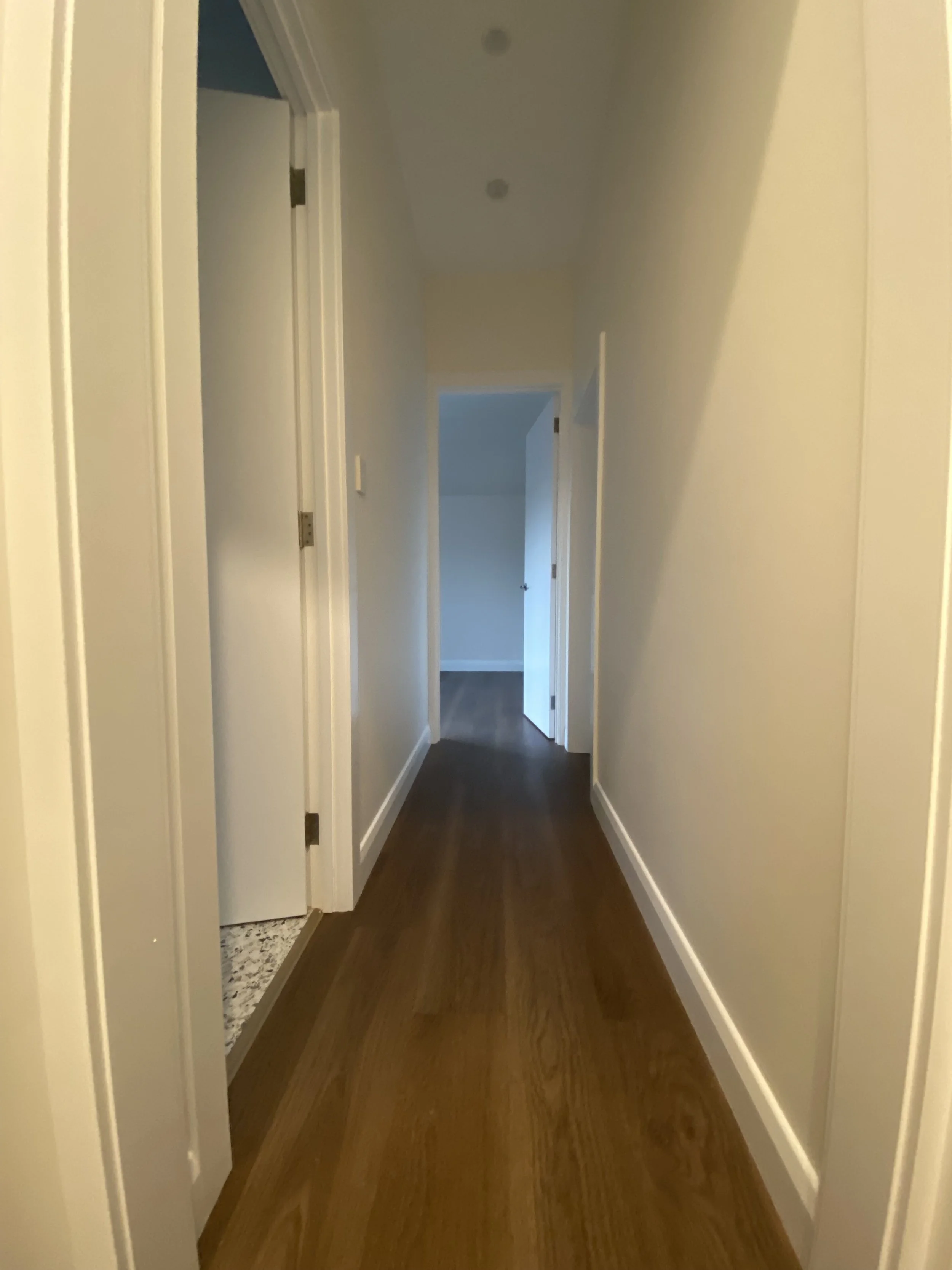 A hallway with wooden flooring, yellow walls, and white door frames leading to multiple rooms. One door is open, revealing a part of a room with a patterned floor and a closed door at the end of the hallway.