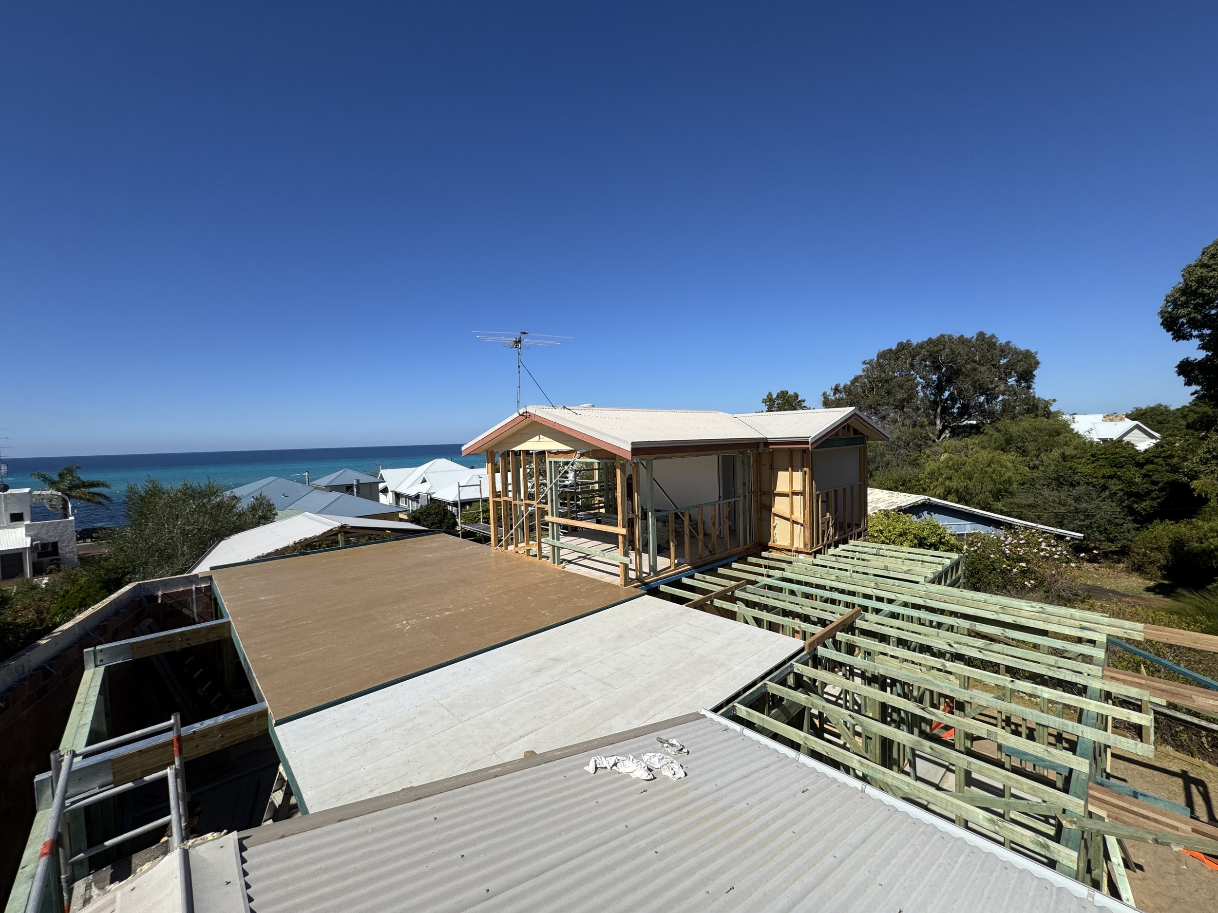 Under construction house with wooden framing, scaffolding, and roofing in a coastal area with blue sky and ocean in the background.