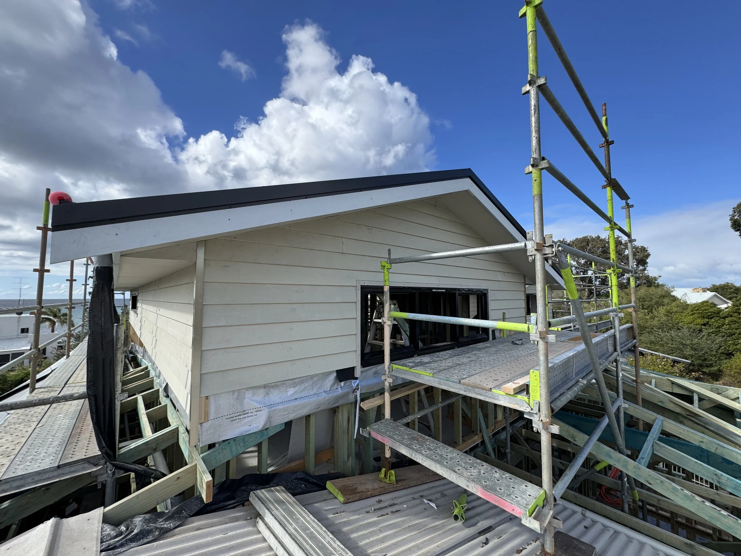 Construction site with a house under construction, scaffolding around the building, and a partly cloudy blue sky.