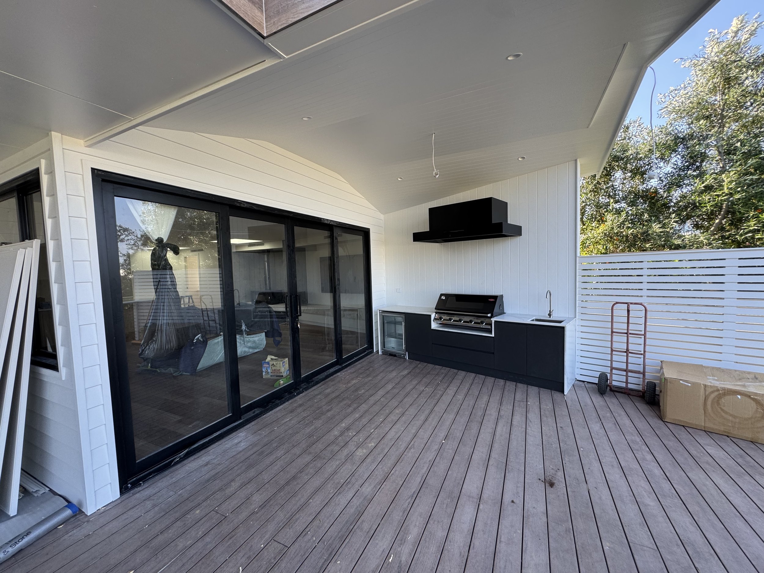 Backyard patio with sliding glass door, black outdoor grill, white walls with black accents, a white fence, and a large green tree. There are some items on the deck, including a portable cart and a cardboard box.