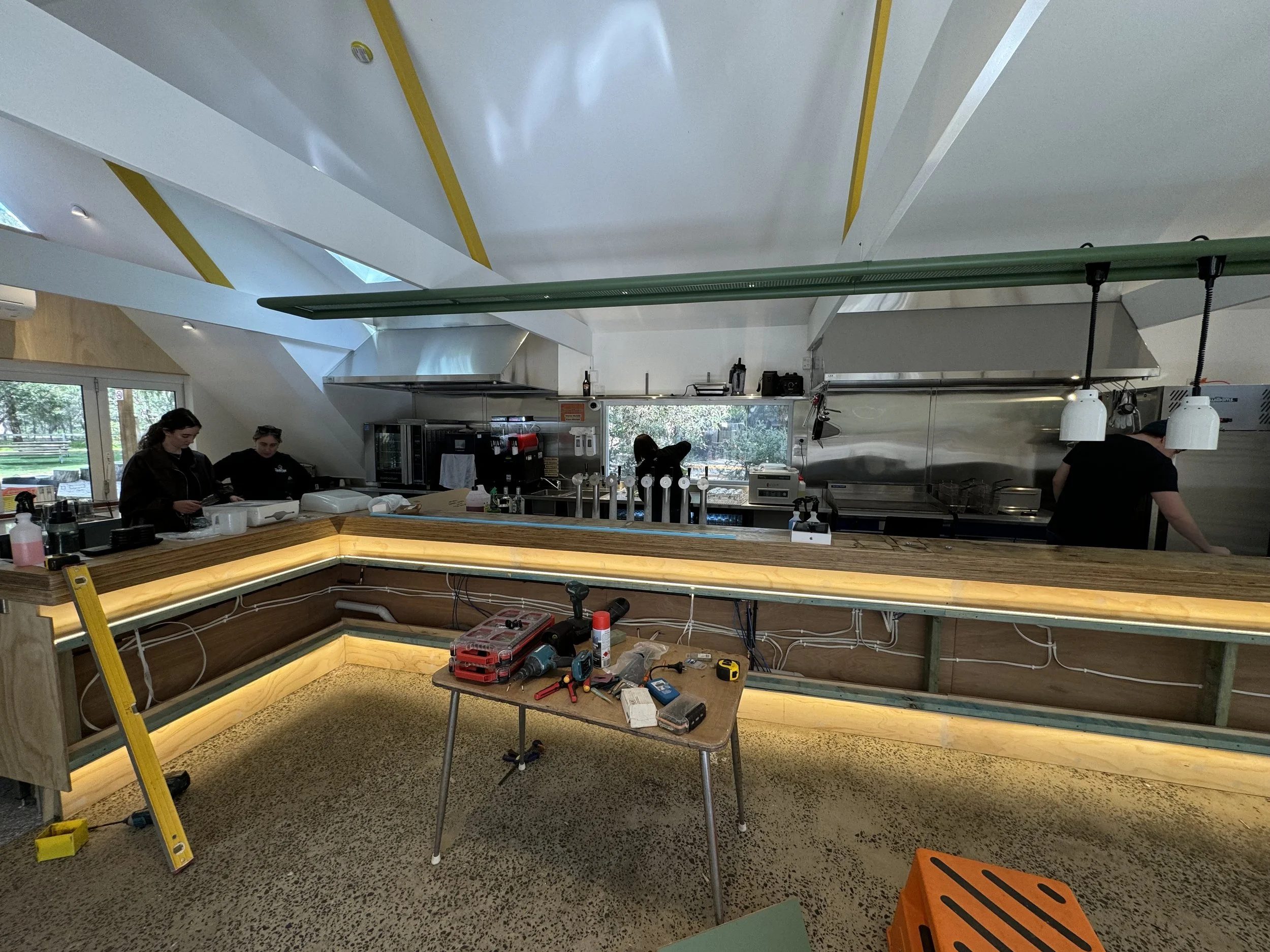 Interior of a restaurant or cafe under construction with workers, tools, and equipment; a partially built wooden counter, tools on a table, and hanging light fixtures.