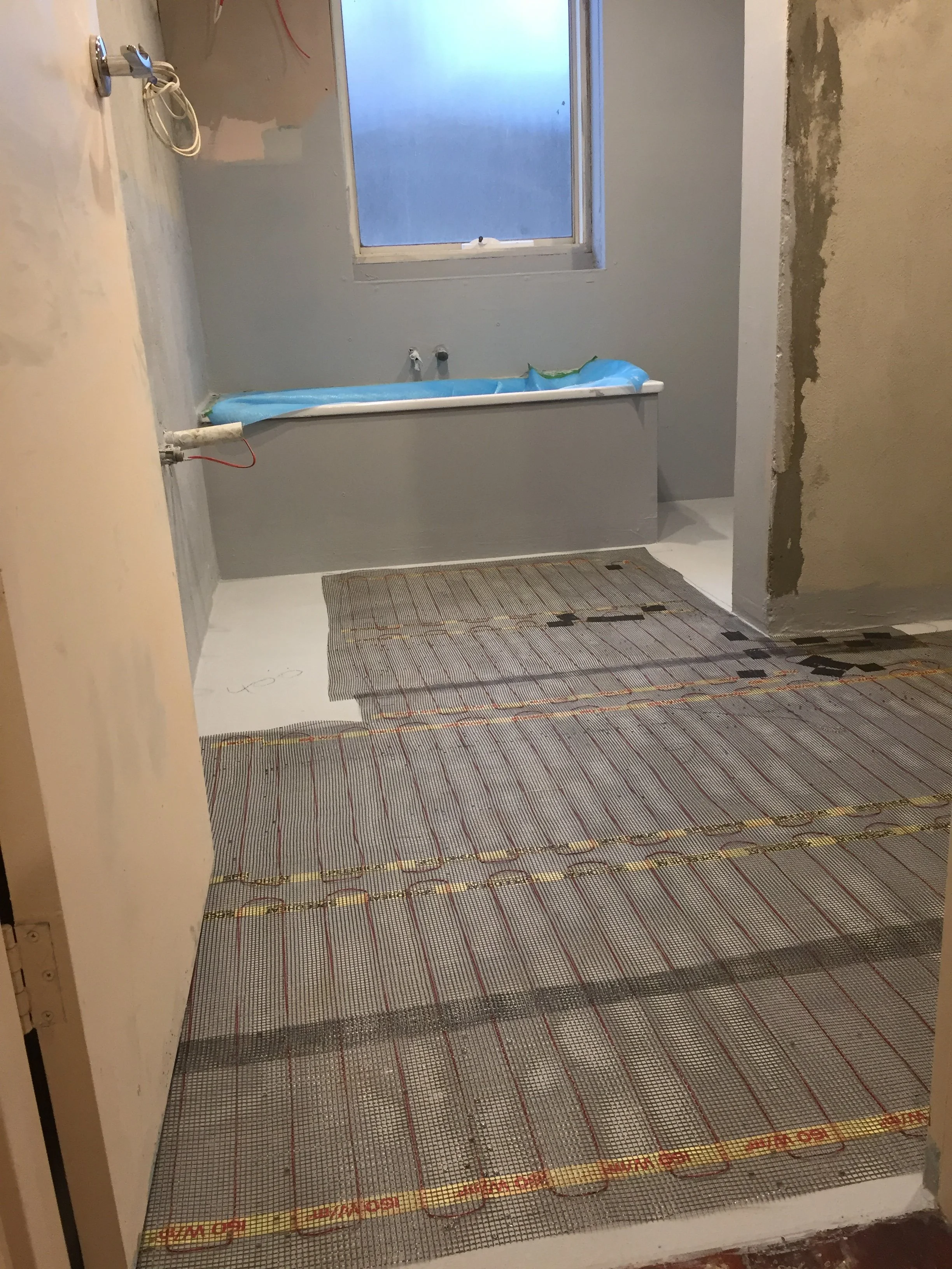 View of a room under renovation with a radiant floor heating system installed on the floor. The room has an unfinished wall on the right side, a window at the back, and a bathtub with blue plastic covering. Construction materials and tools are presen