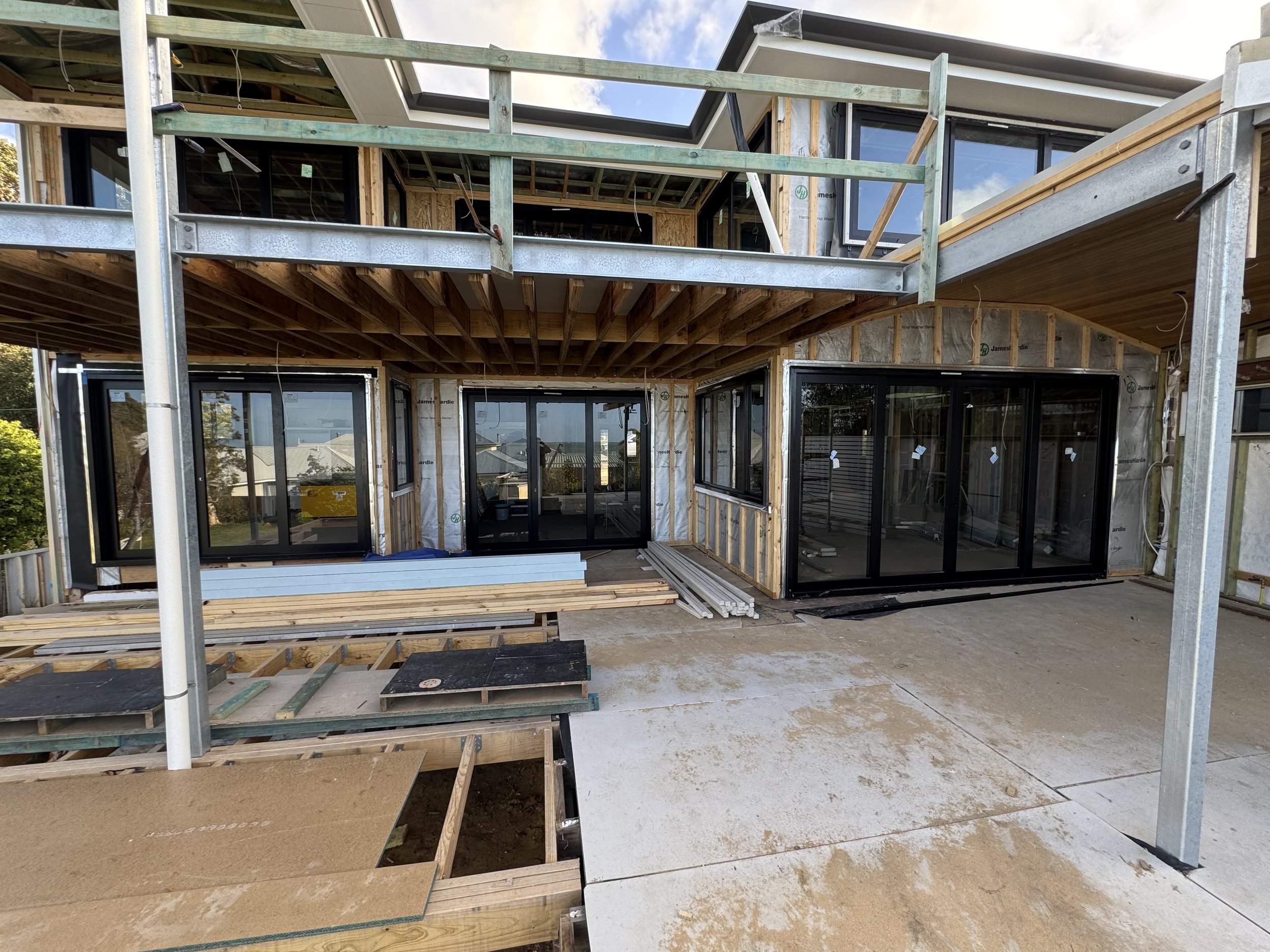 Construction site of a two-story house with scaffolding, windows, and exposed wooden framing under a partly cloudy sky.