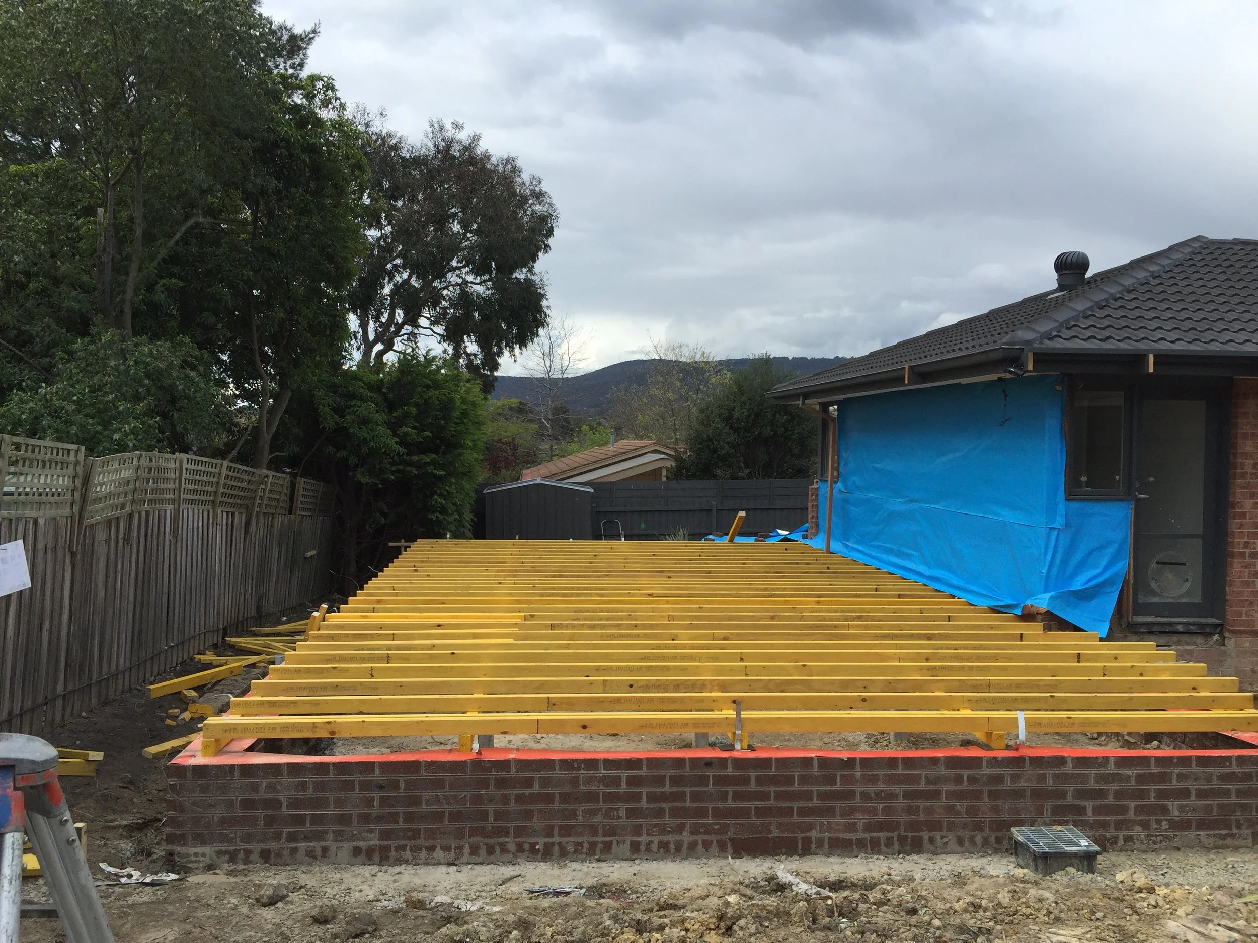 Construction site with wooden frames for a decking area, a house with a blue tarp covering part of the exterior, trees, cloudy sky, and distant hills.
