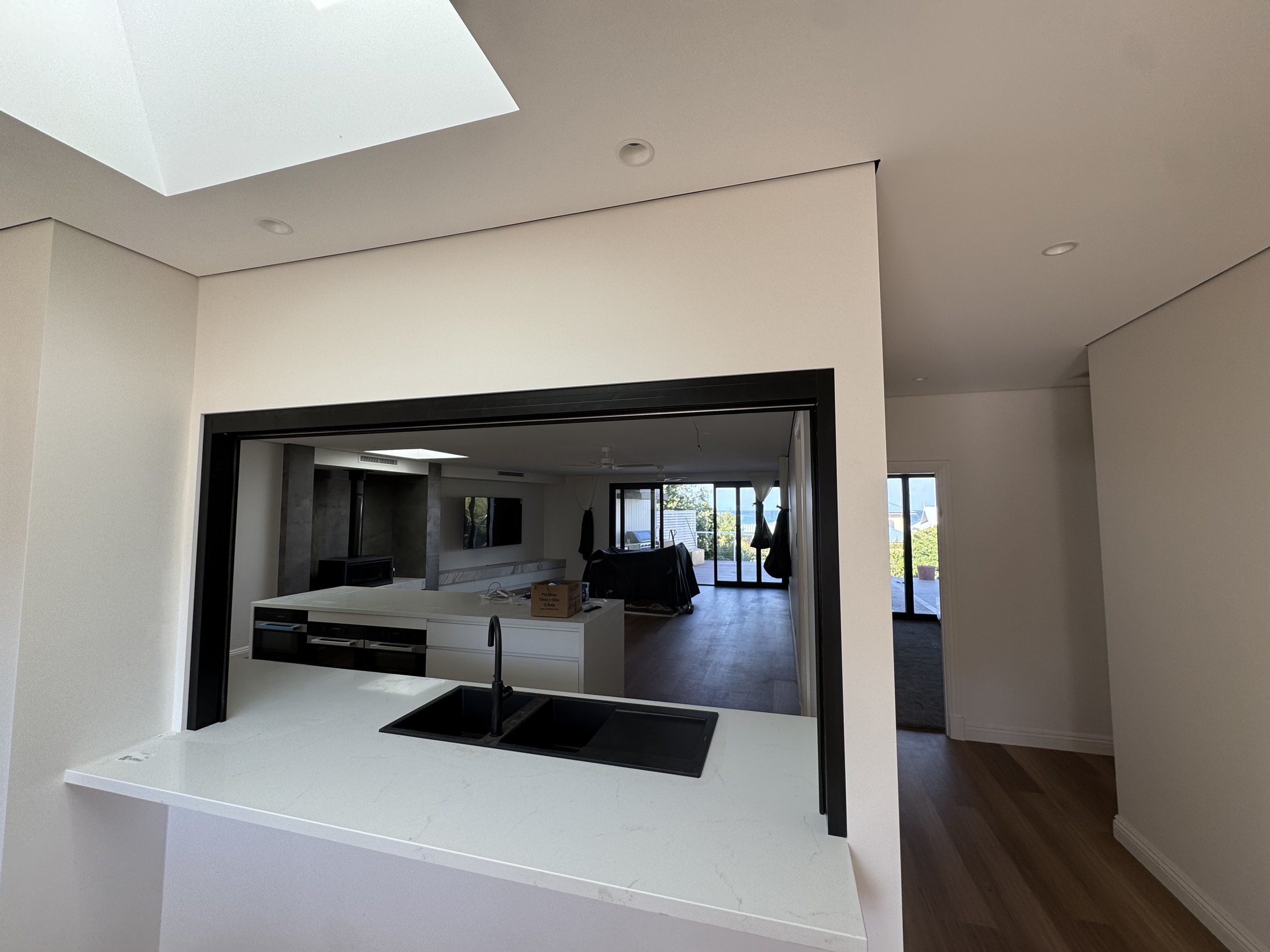 Modern open-concept kitchen with a white island, black sink, and faucet, overlooking a living area with large windows and sliding glass doors to an outdoor space.