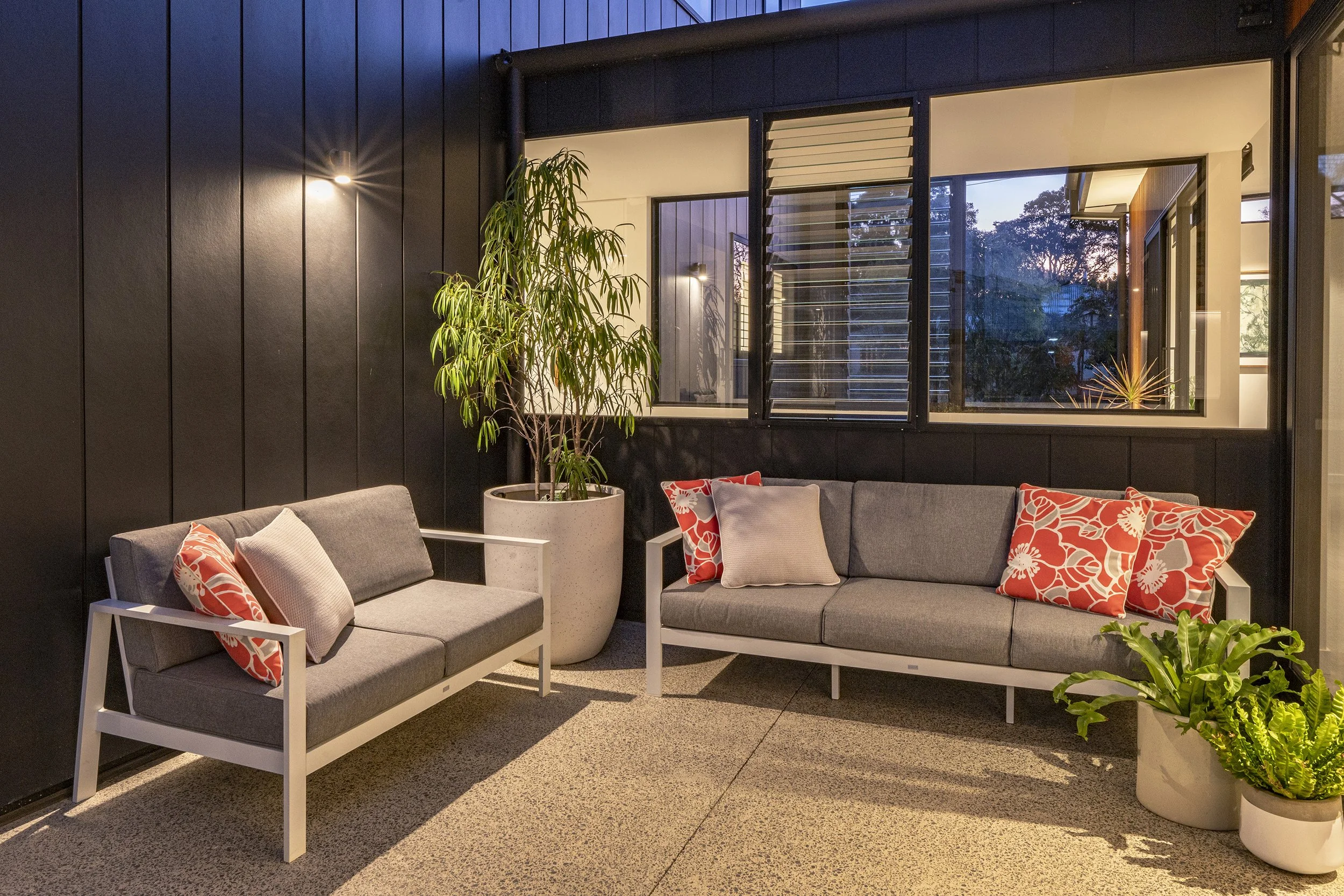 A cozy indoor seating area featuring two matching gray sofas with white frame, colorful red and white floral pillows, a potted plant, and large windows showing trees outside at dusk, with dark walls and warm lighting.