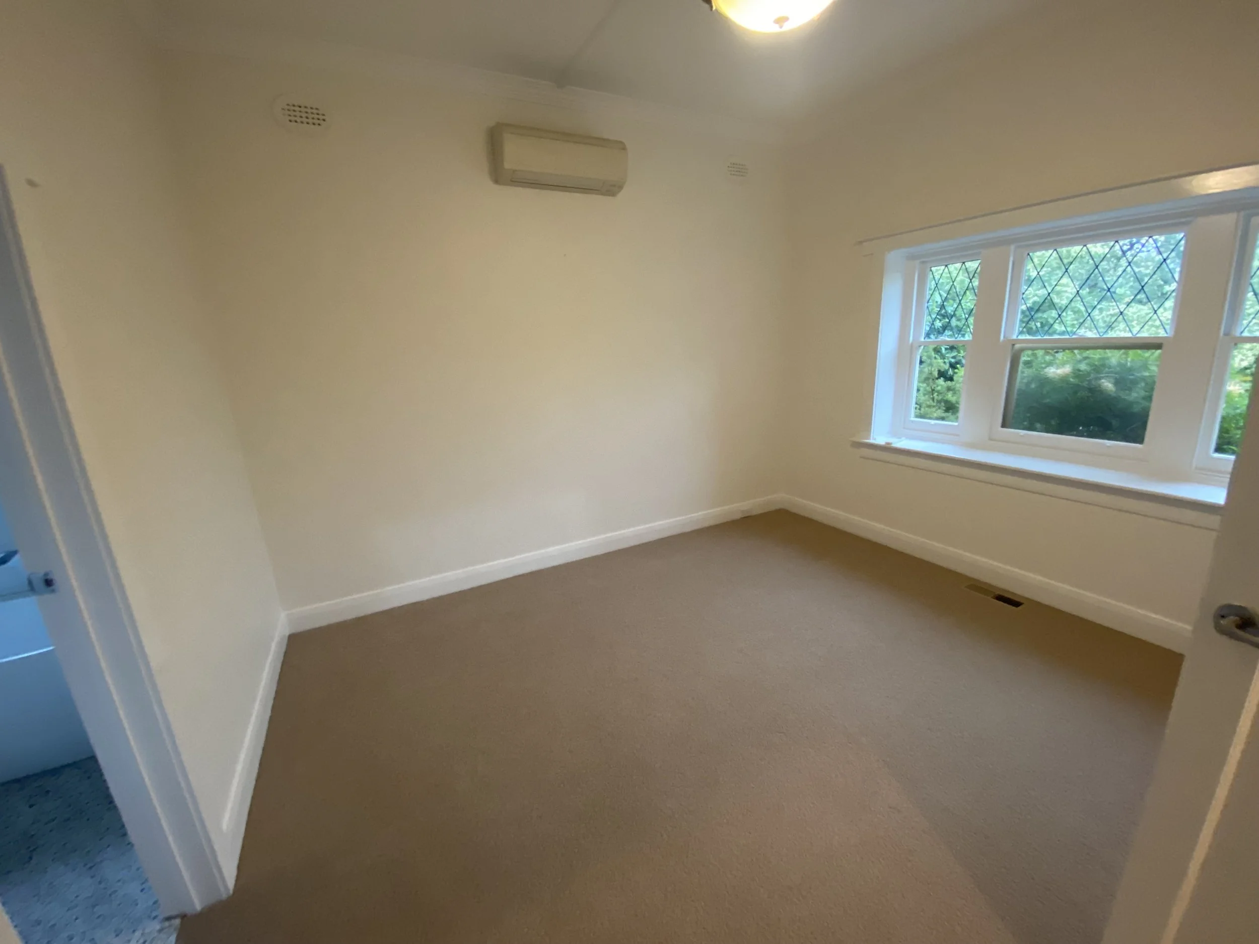Empty beige carpeted room with white trim, window with diamond grid pattern, and a white air conditioning unit on the wall.