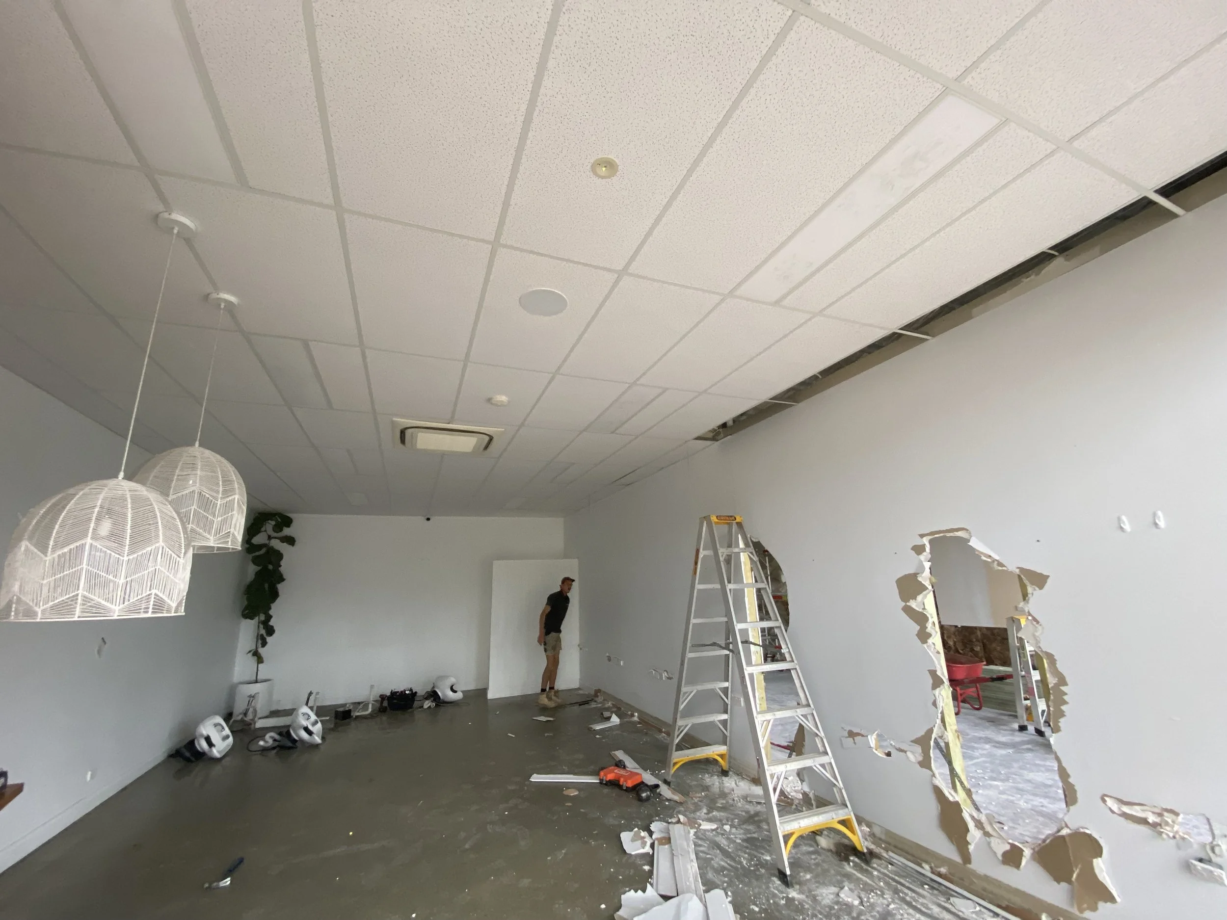 A room under renovation with a damaged ceiling and construction tools. Two white pendant lights hang from the ceiling, and a ladder is set up in the middle of the room. There are some debris and a person working on the wall.
