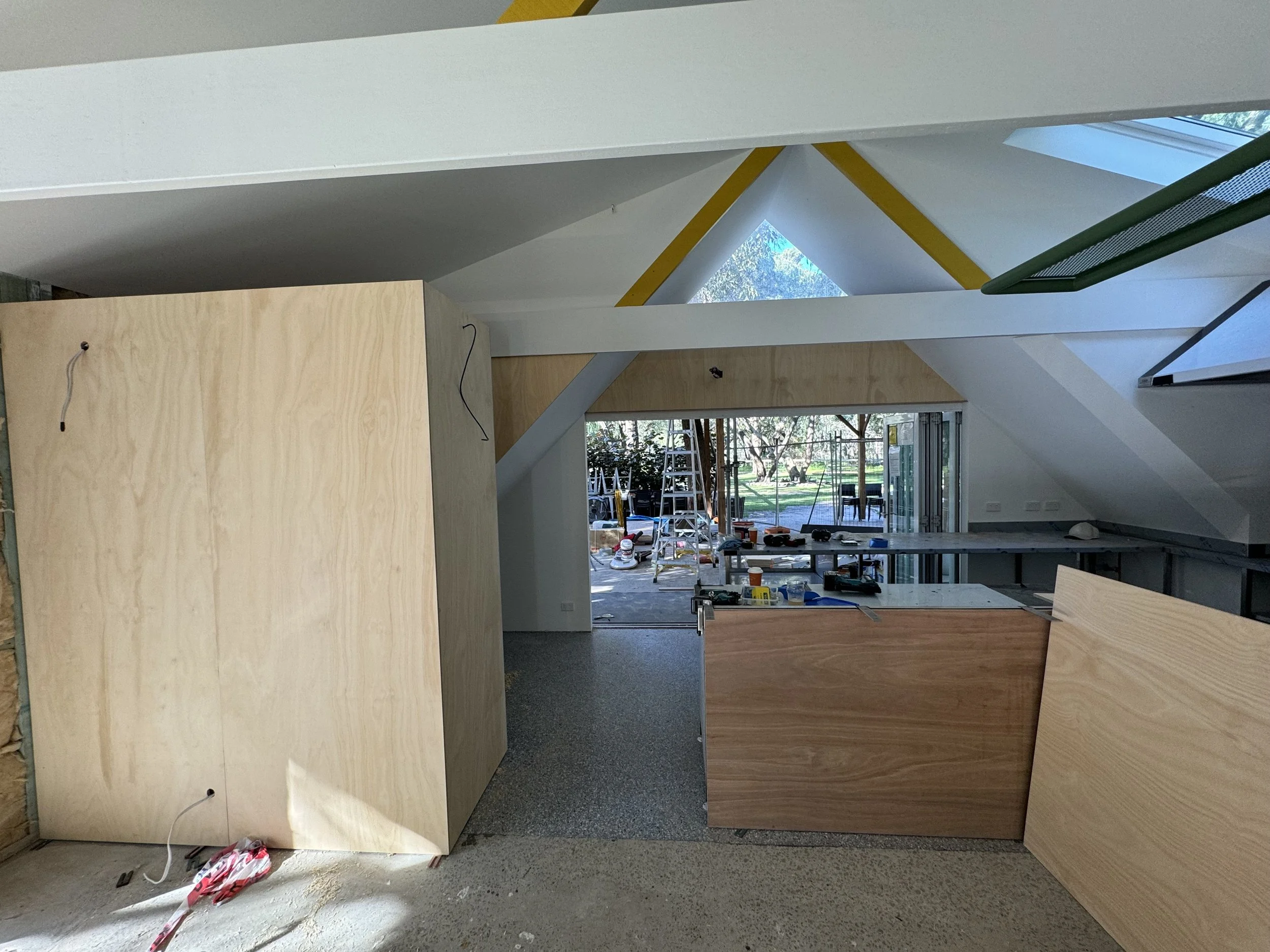 Interior of a building under construction with plywood panels, construction tools, and large windows showing trees outside.