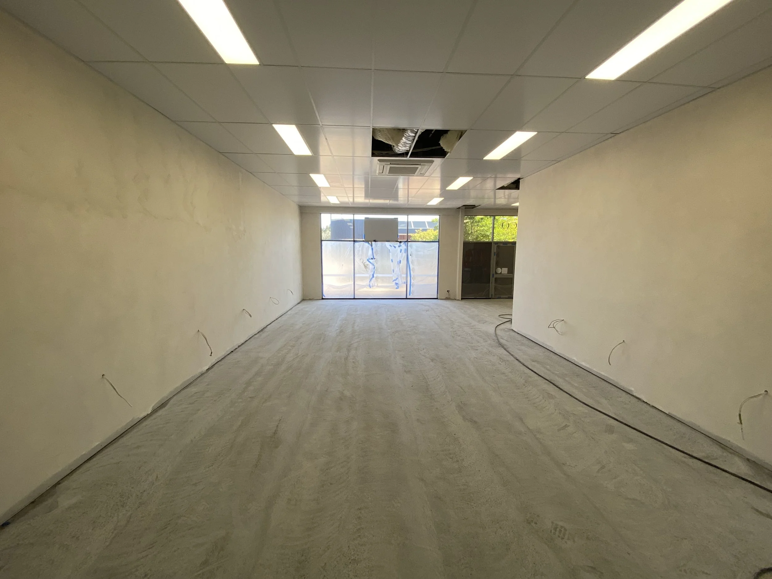 An interior view of an unfinished room with a boarded-up entrance and open ceiling panels showing ductwork, with a window at the back letting in natural light.
