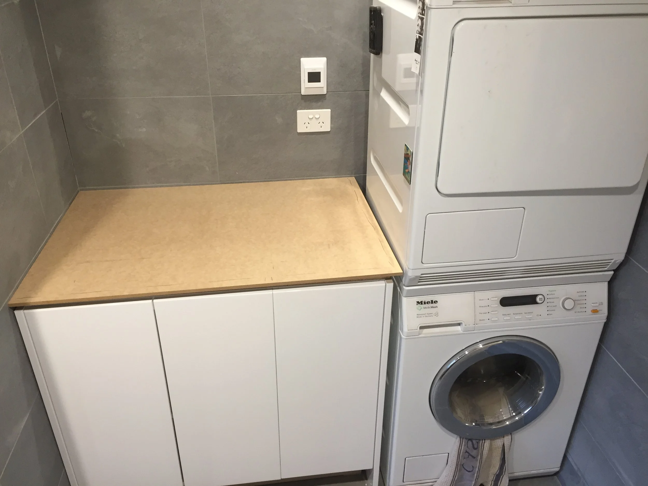 A laundry room with gray tiled walls and floor, featuring a white washing machine and dryer positioned side by side, with a wooden shelf mounted on the wall above them.