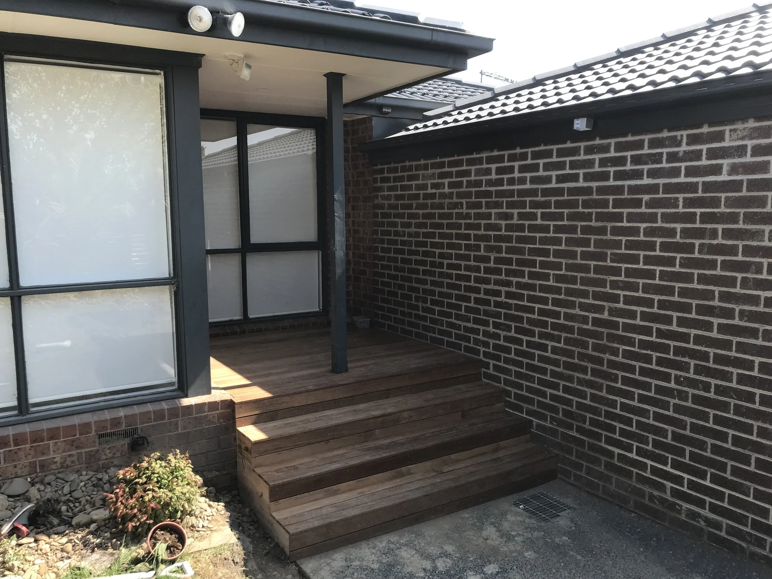 A small outdoor area with a wooden deck, brick step, black brick wall, and sliding glass doors leading into a house. There are two small outdoor lights on the ceiling and one on the brick wall.