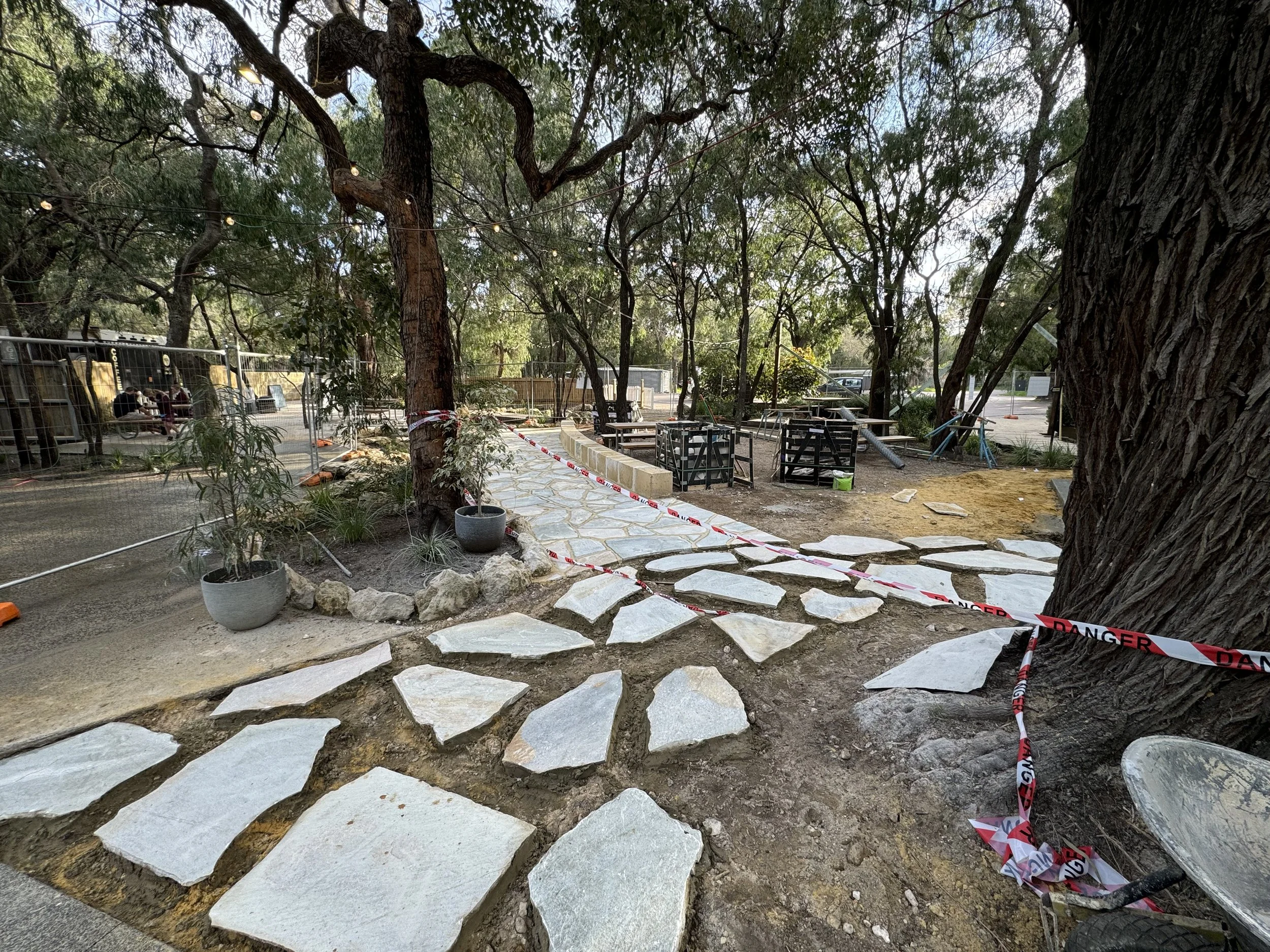 Construction site with stone walkway, trees with string lights, and fencing, indicating ongoing landscaping or outdoor project.