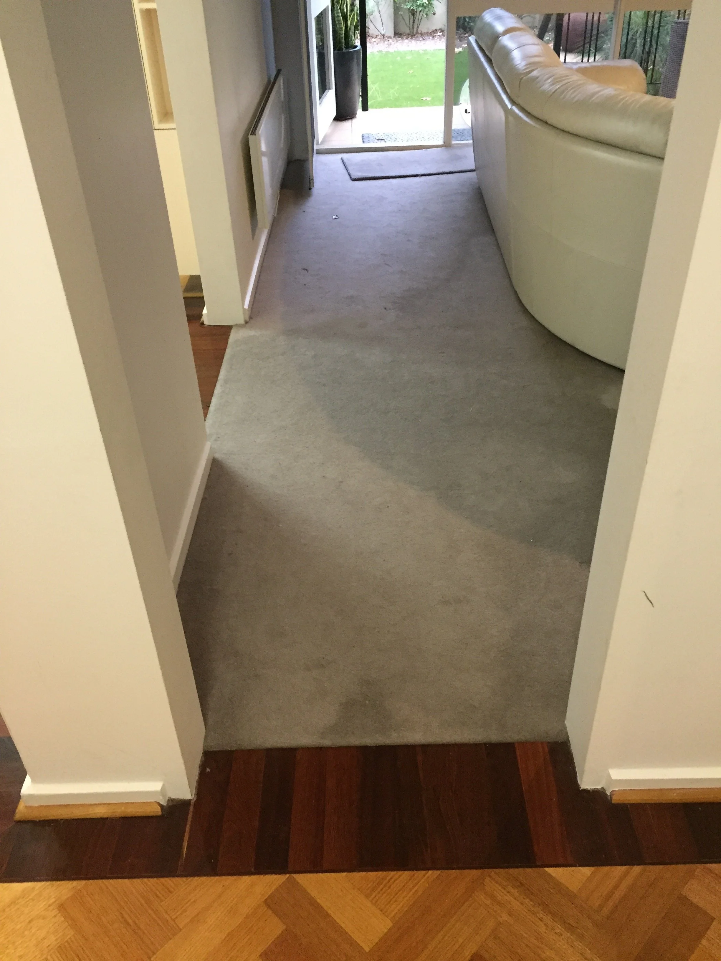View from inside a house showing a hallway with a beige carpet leading to a sliding glass door that opens to a backyard with green grass and plants. There is a white leather couch on the right side and a black mat in front of the door.