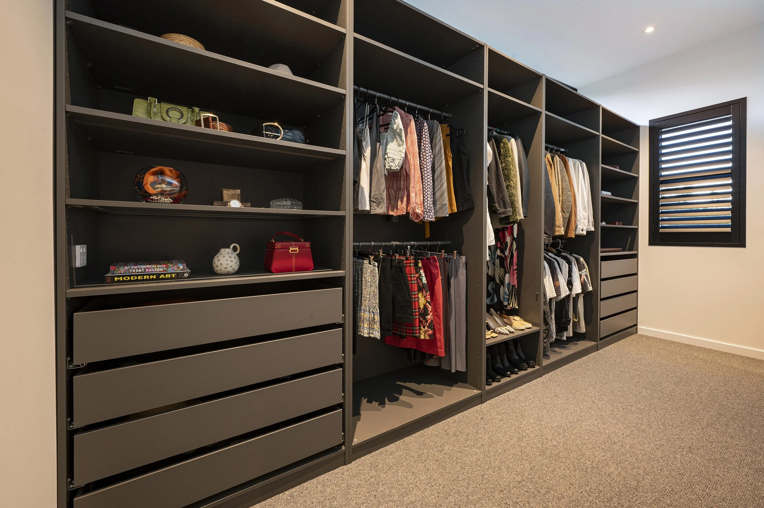 A walk-in closet with black shelving units filled with hanging clothes, shoes, and accessories, and a window with black shutters on the right side.